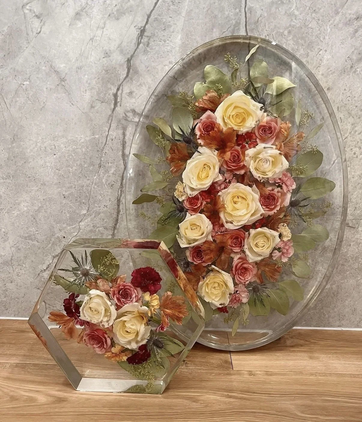 A decorative floral arrangement with preserved roses and greenery in a large oval glass tray, alongside a smaller glass container with similar flowers, placed on a wooden surface against a stone wall background.
