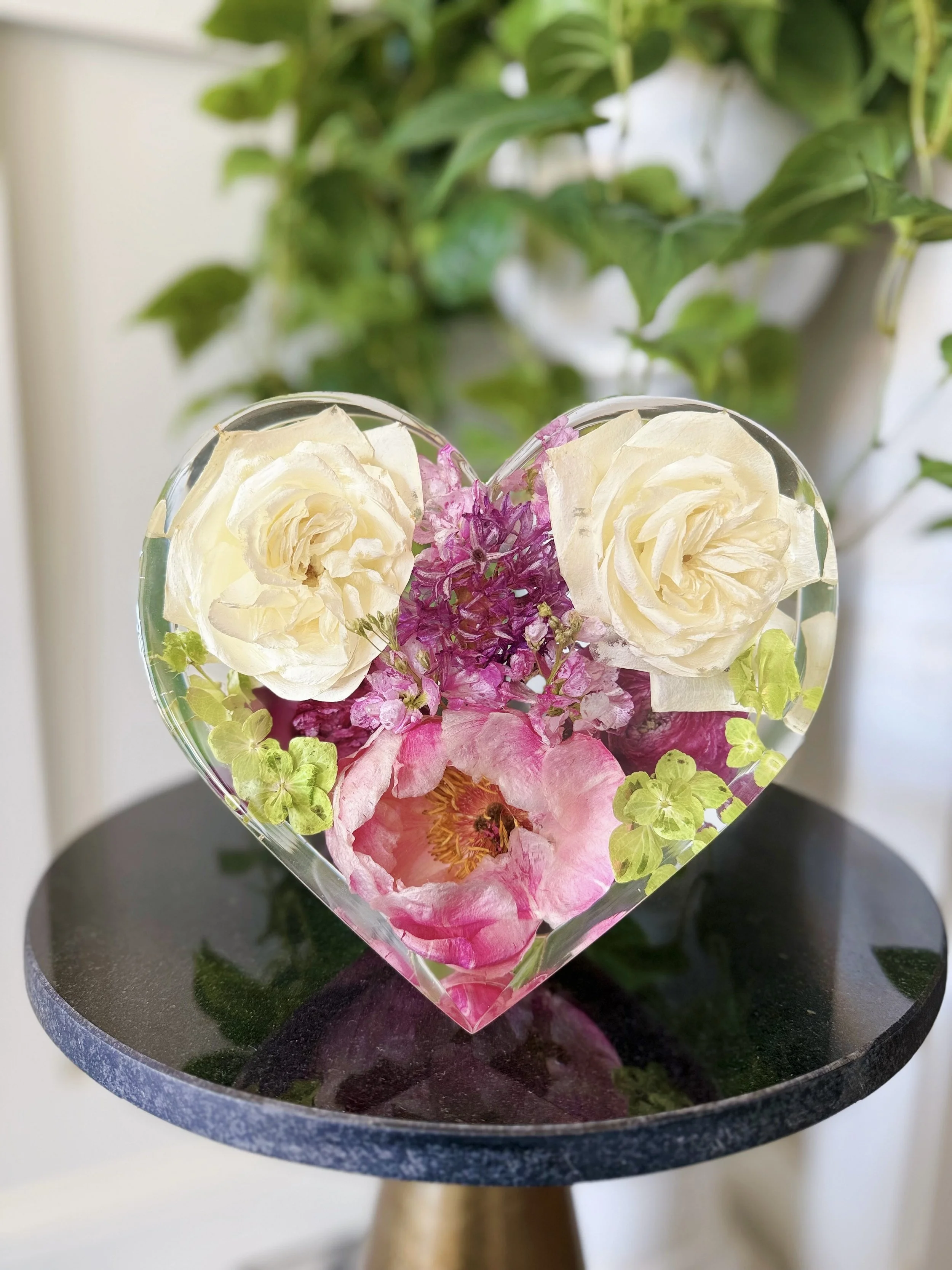 A heart-shaped resin mold filled with white roses, pink flowers, and green foliage, placed on a dark round tabletop with a green plant in the background.