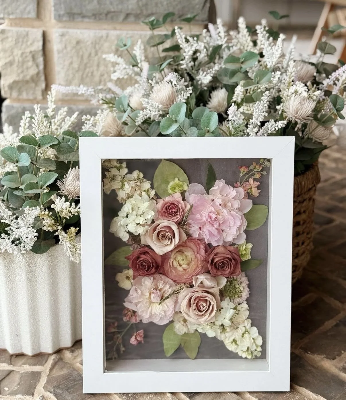 A framed floral display featuring pink and white roses, hydrangeas, and various greenery, placed against a background of additional white flowers and a basket of flowers.