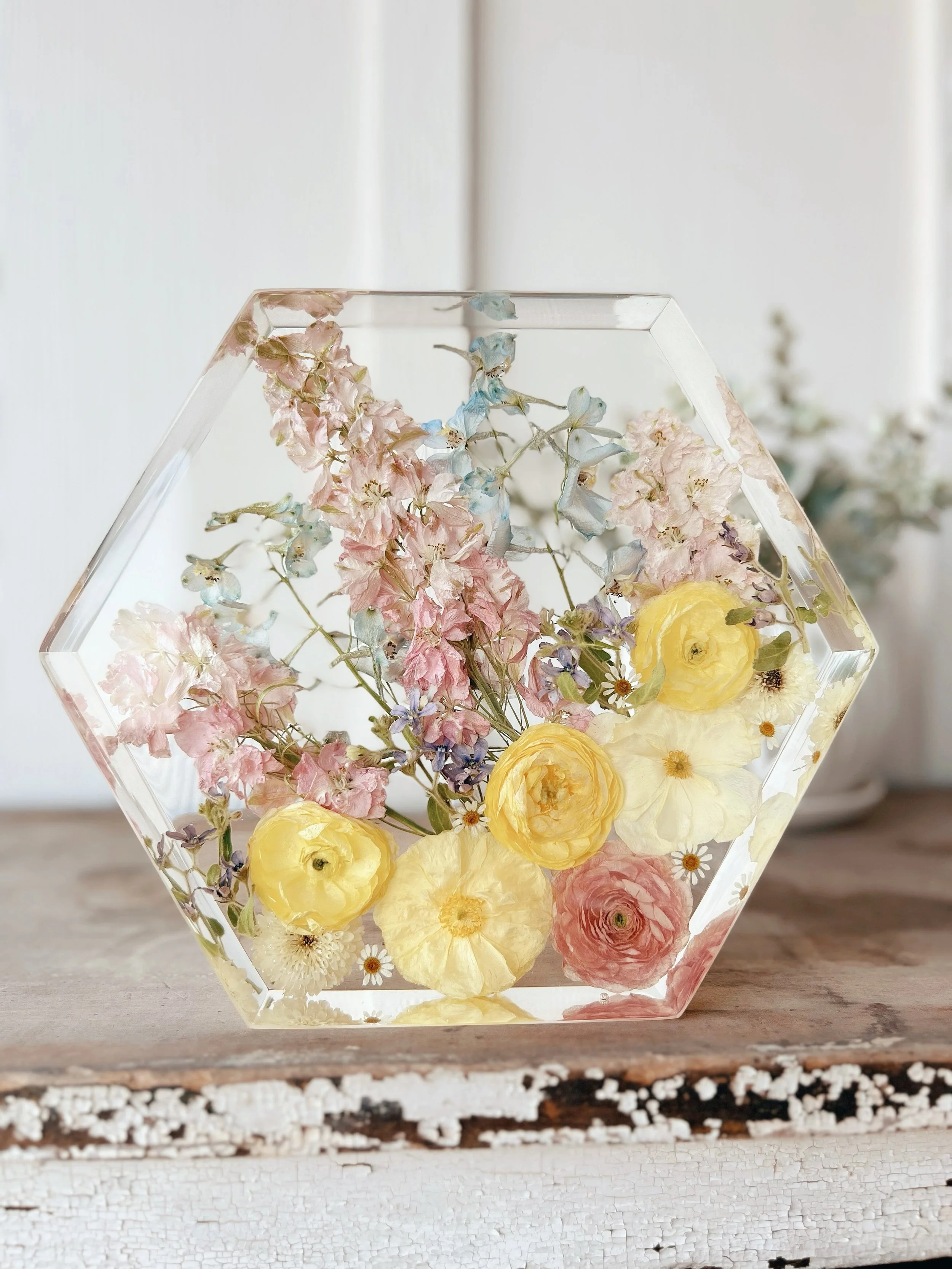 A hexagonal glass vase filled with water and various pastel-colored flowers, including pink, yellow, and blue flowers, sitting on a rustic wooden surface with a blurred background of white and green.
