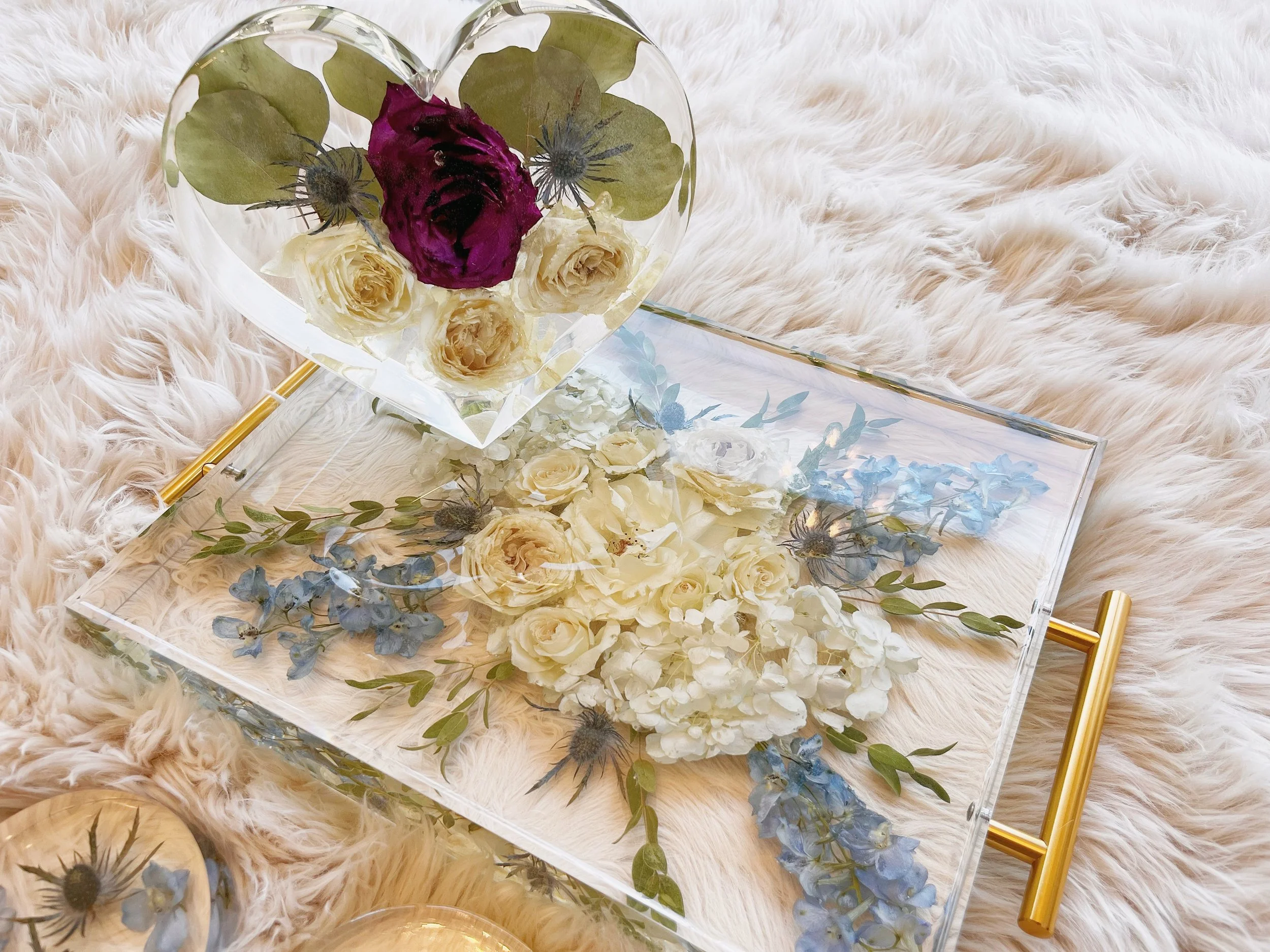 A clear tray with floral arrangements and a heart-shaped glass container with pressed flowers inside. The tray is placed on a soft, cream-colored furry surface.