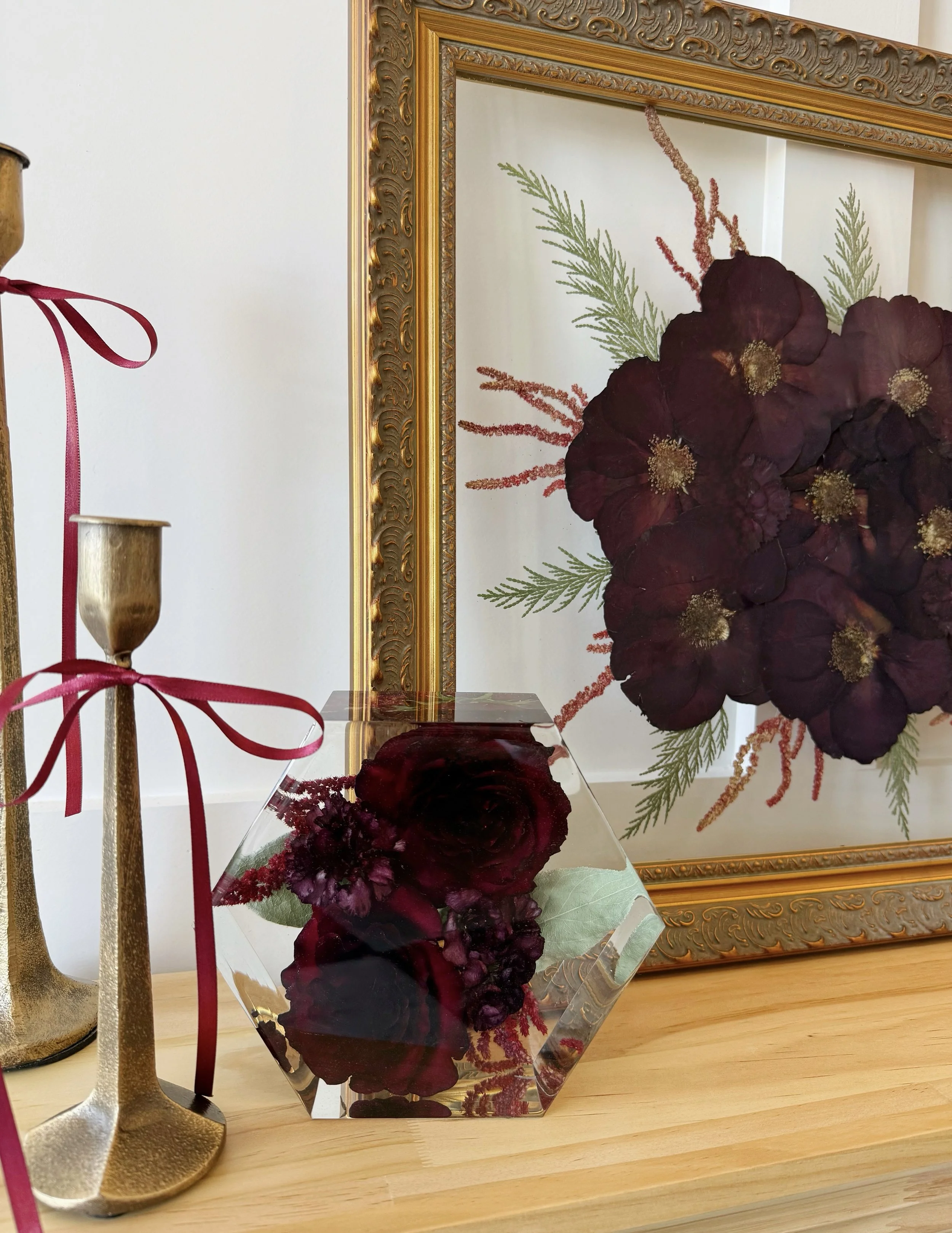 Decorative display featuring a framed floral artwork with dark purple flowers and green foliage, a glass cube containing a dark purple flower and greenery, and tall gold candlesticks tied with red ribbons on a wooden surface.