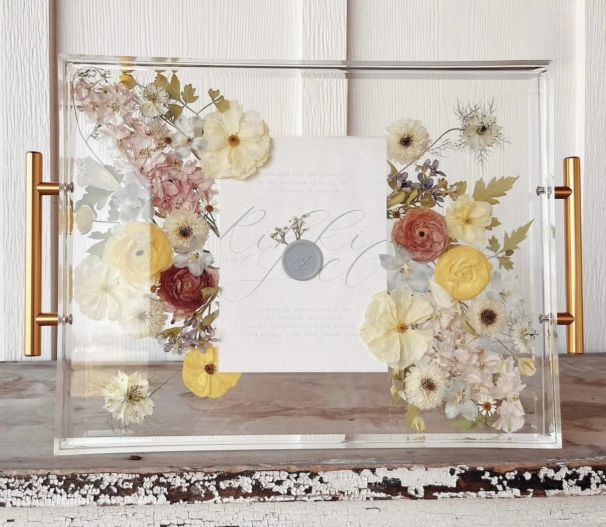 A transparent display case with gold handles filled with mixed dried flowers and a white document inside.