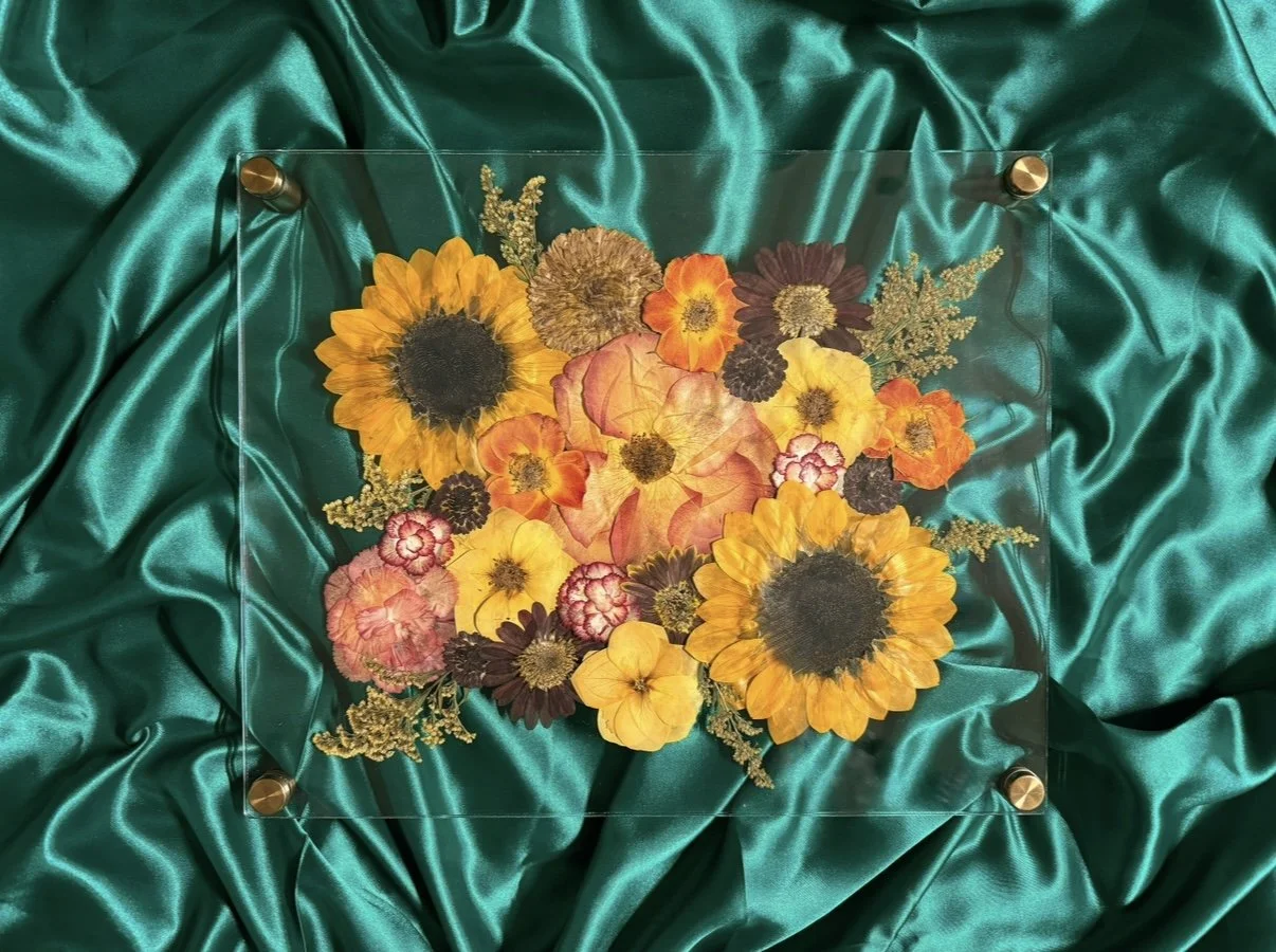 Pressed flower arrangement with sunflowers, marigolds, and other flowers on teal satin fabric.