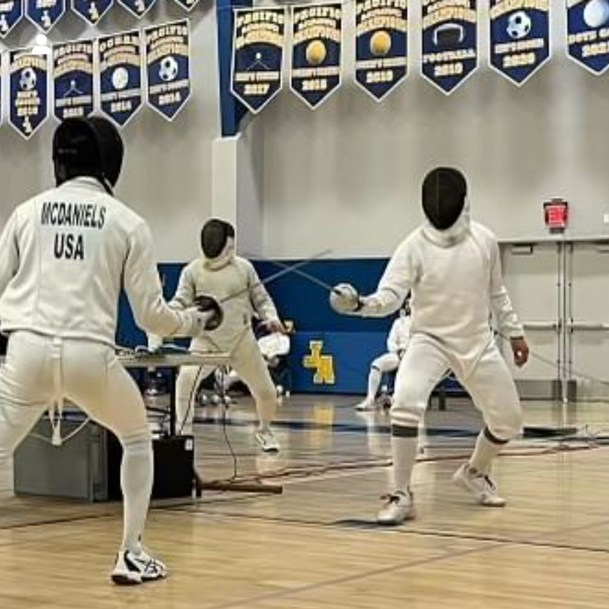 Two men fencing with swords in a gymnasium, wearing white protective gear, overlooking banners and a basketball court.