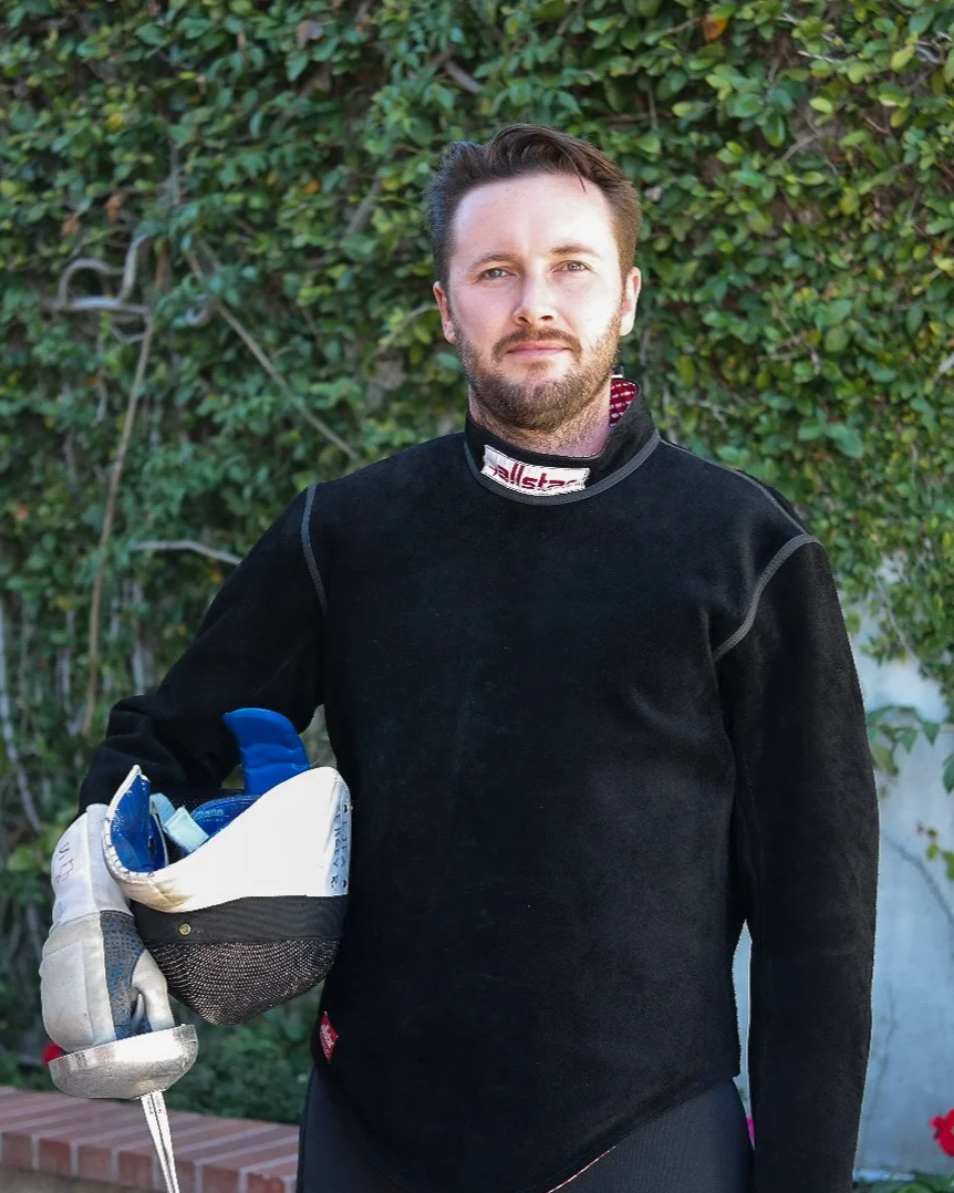 Man in black racing suit holding a fencing mask with fencing gear inside, standing outside with green foliage in the background.