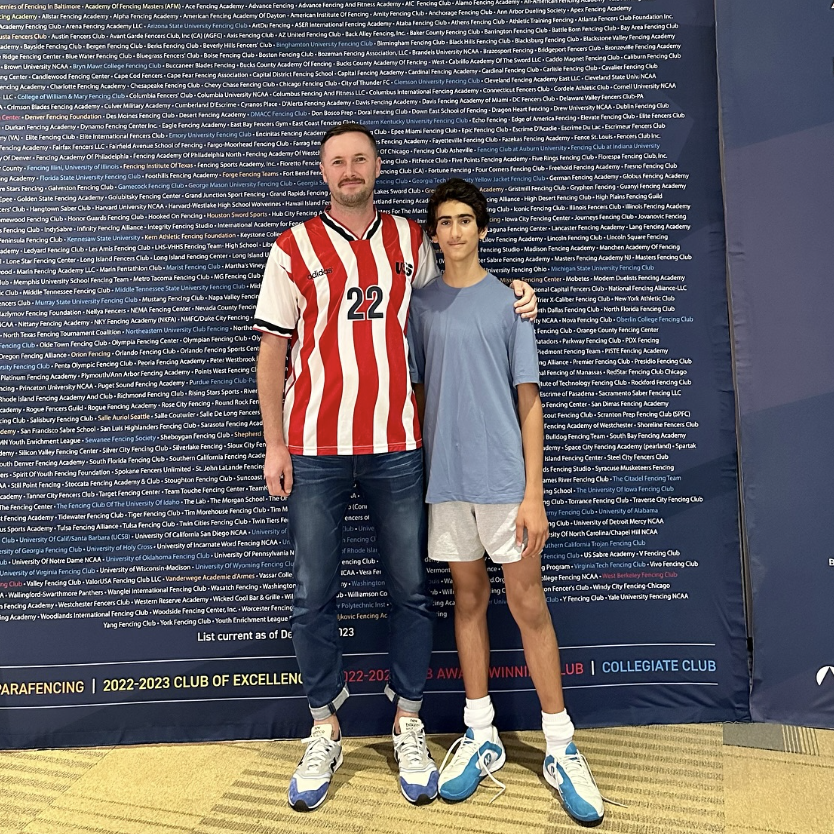 Two young males standing together, one in a red and white striped hockey jersey with the number 22, and the other in a light blue T-shirt and gray shorts, at a fencing club event. Both are wearing sneakers and have their arms around each other. The b