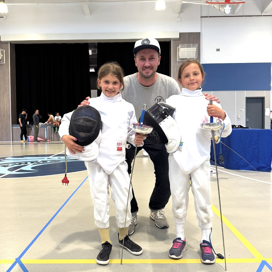 Two young girls in fencing uniforms holding fencing masks and swords, posing with an adult man in a gymnasium or sports hall, who is smiling and standing behind them. The girls are smiling and holding their fencing gear, with the background showing o