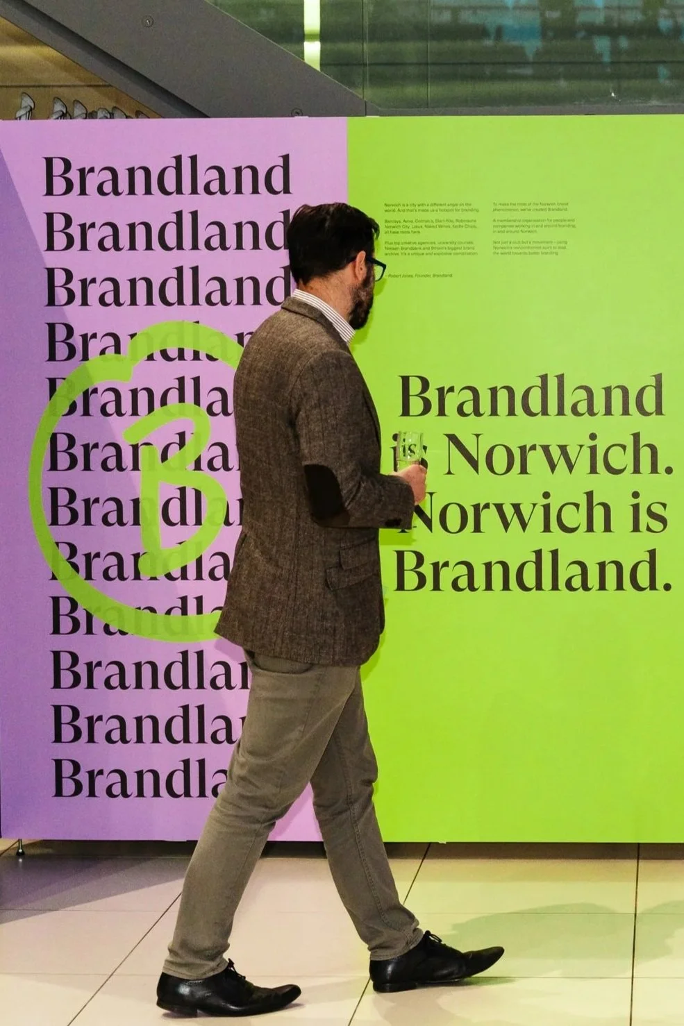 A man with glasses, a beard, wearing a brown blazer, striped shirt, gray pants, and black shoes is standing in front of a colorful display that reads "Brandland, Norwich." The display has a pink and green background with repetitive text and a large question mark.