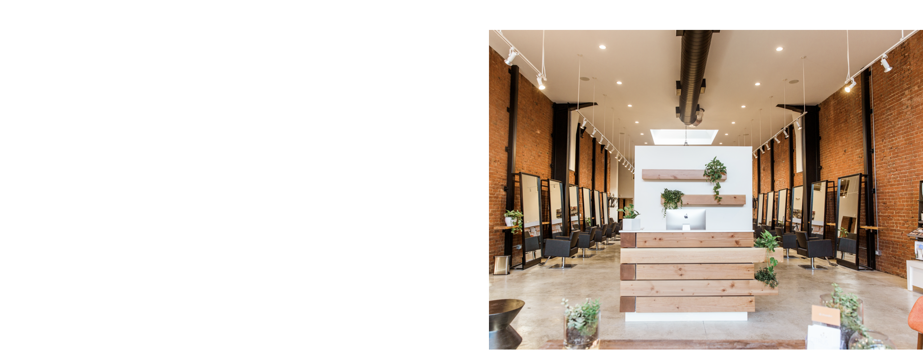 Modern hair salon with brick walls, several styling stations with mirrors and chairs, and a reception desk with wooden panels and plants.
