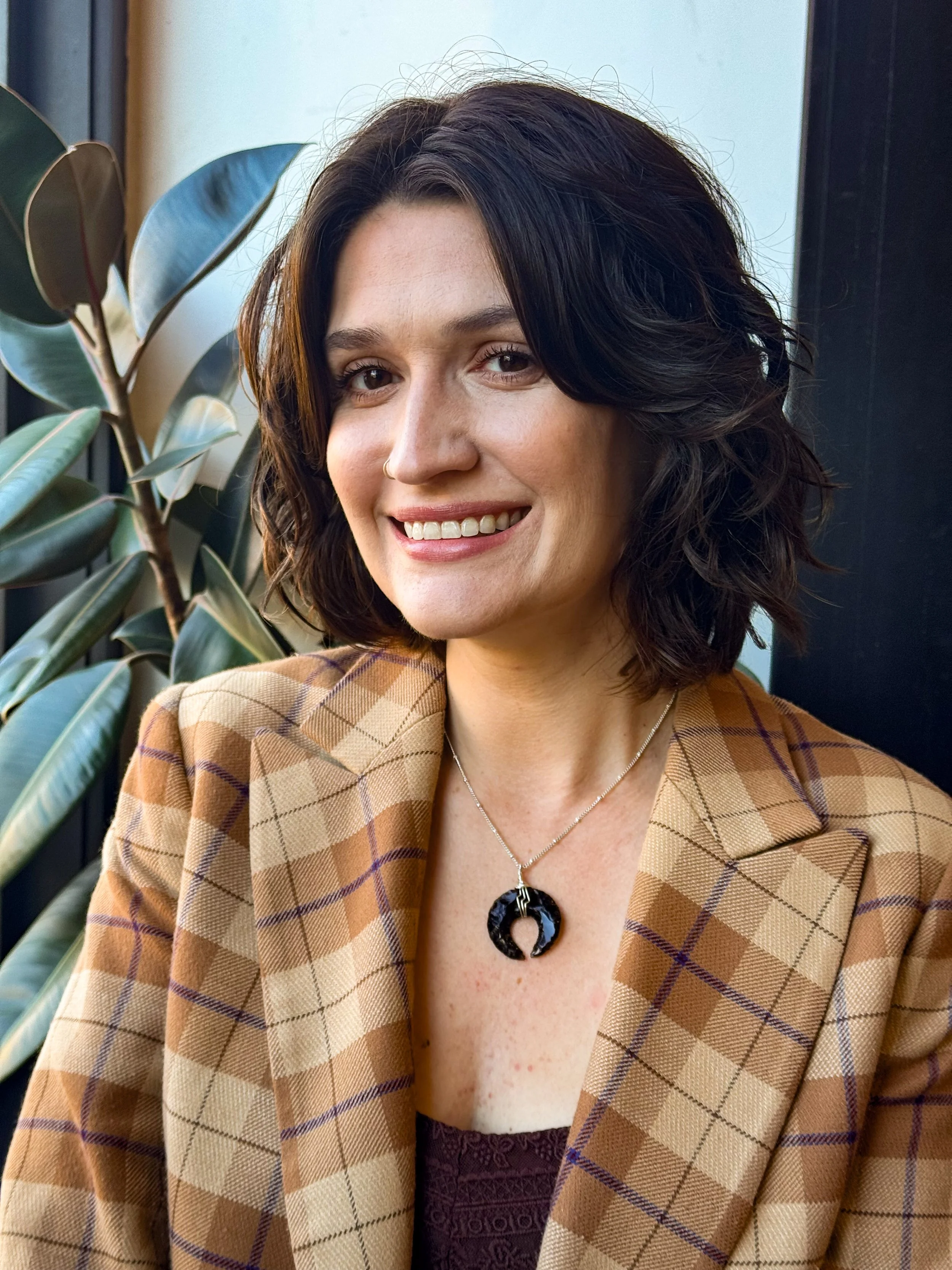 Close-up of a woman with dark, wavy hair, smiling and wearing a brown checked blazer, a black necklace with a circular pendant, and a purple patterned top, standing by a window with green plants.