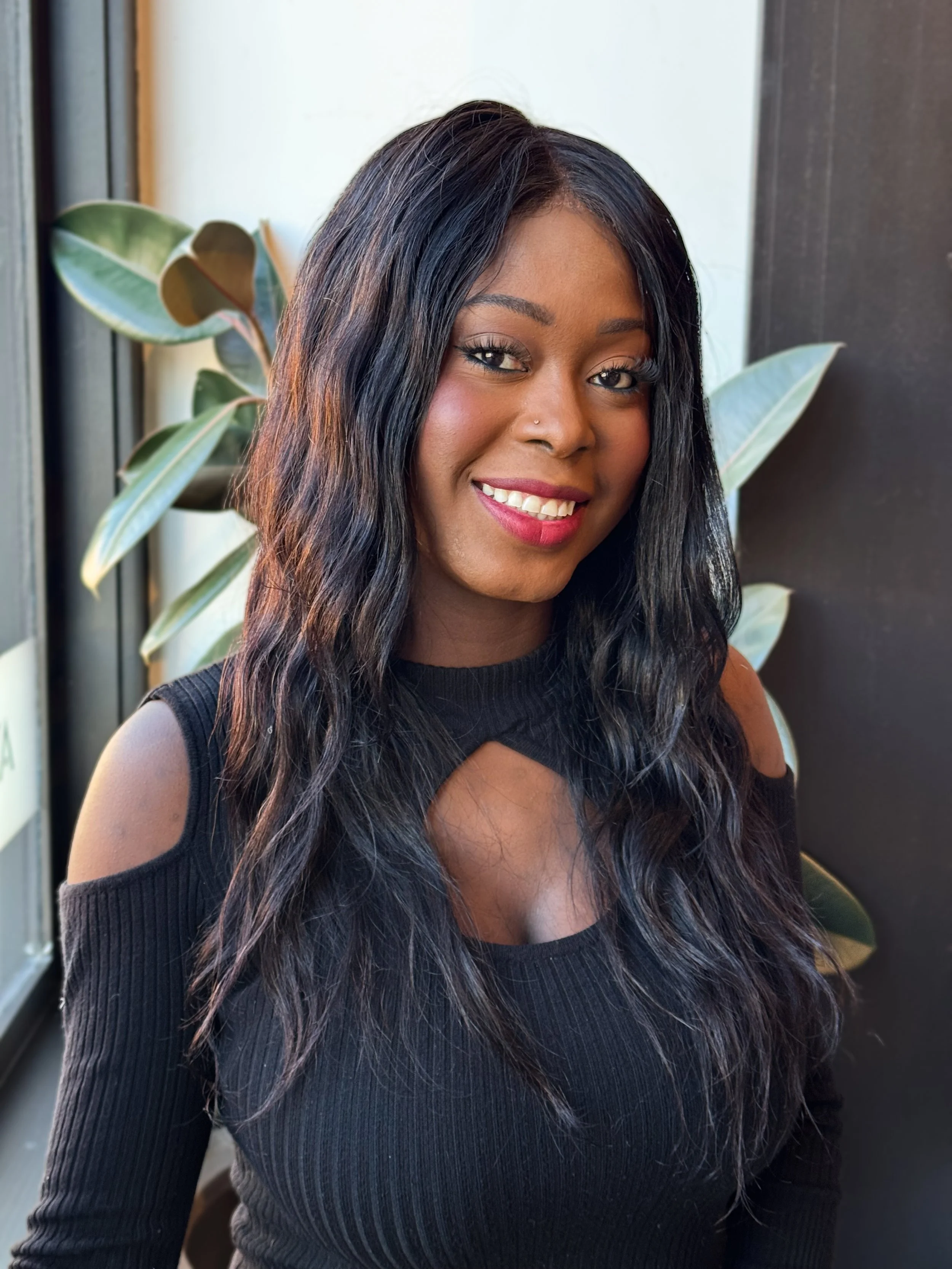 A woman with long dark wavy hair smiling, wearing a black cut-out top, standing next to a plant in an indoor setting.