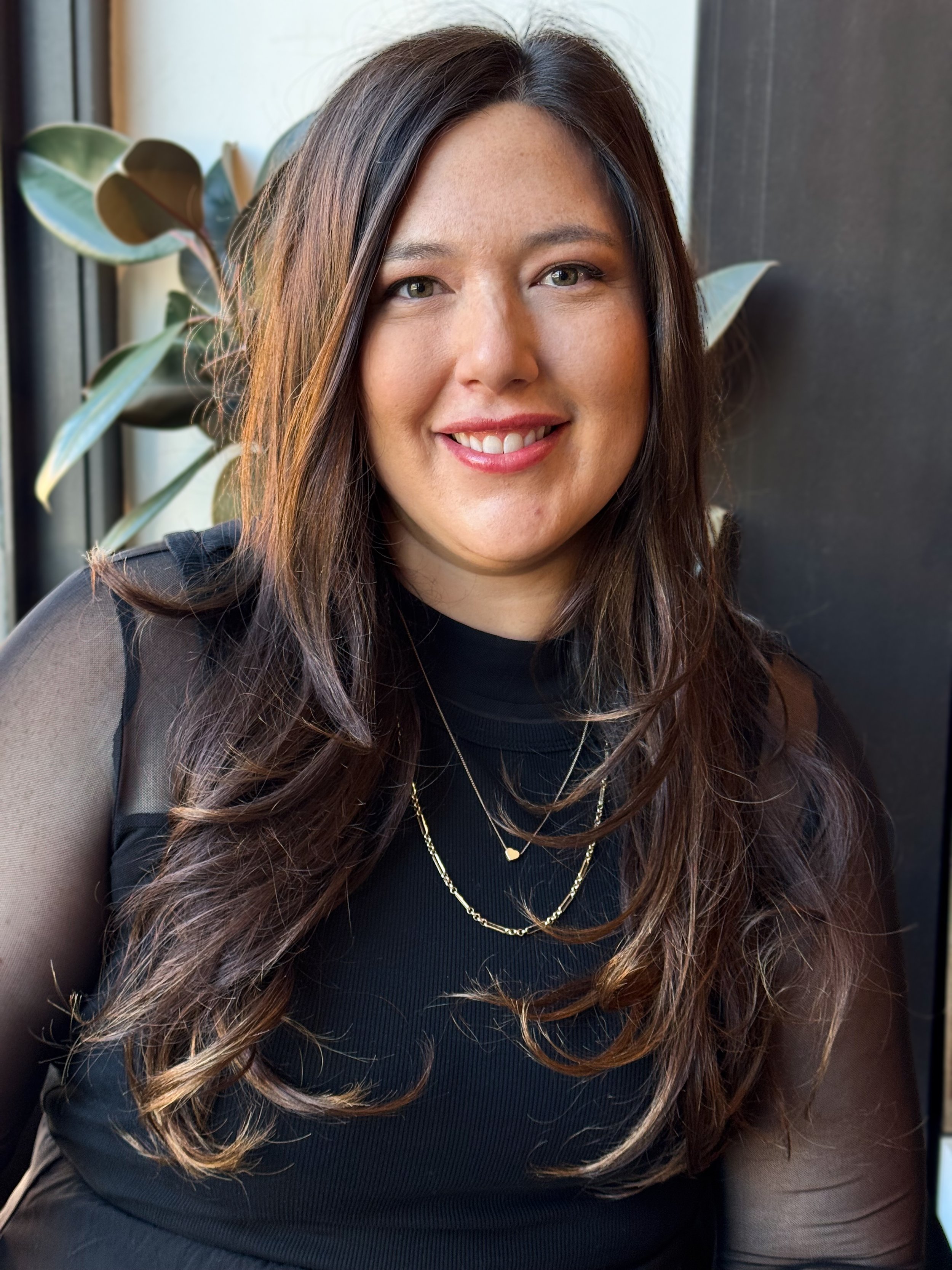 A young woman with shoulder-length dark brown hair and light skin, smiling, wearing gold hoop earrings, a nose ring, and a black ribbed top with ribbon ties at the neckline, sitting indoors near a window with green plants in the background.