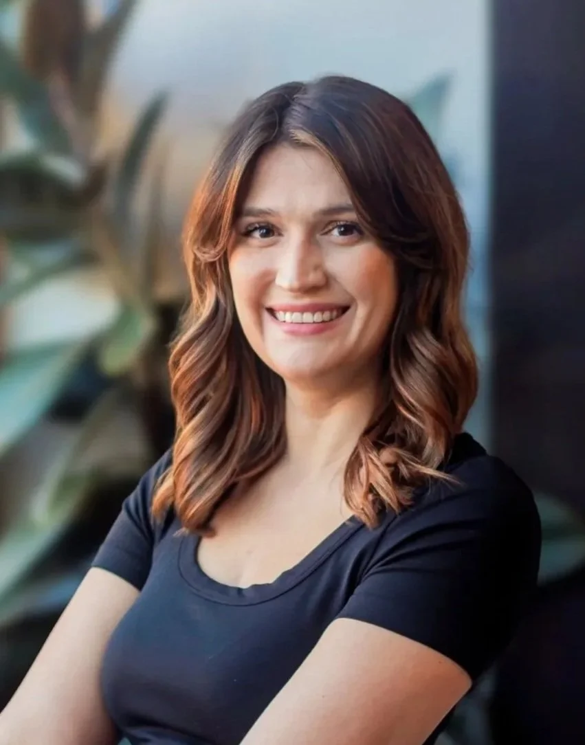 Portrait of a woman with shoulder-length wavy brown hair, smiling, wearing a black blouse and a silver chain necklace, with a plant and window in the background.