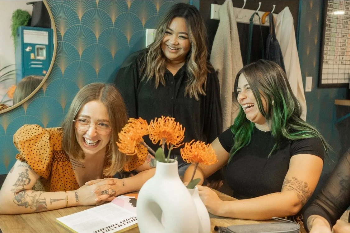 Three women sitting at a table, laughing and enjoying each other's company. One woman with blonde hair and glasses, another with dark-green streaked hair, and a third standing behind them with wavy hair. There is a white vase with orange flowers on the table, and a mirror on the wall behind them.