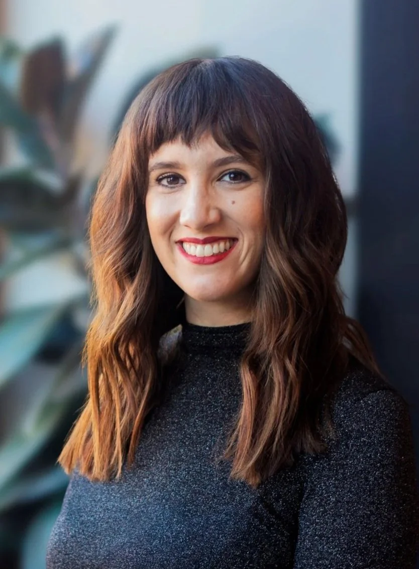 A woman with long wavy brown hair, red lipstick, and a nose piercing, wearing a black top, looking to the side and smiling.