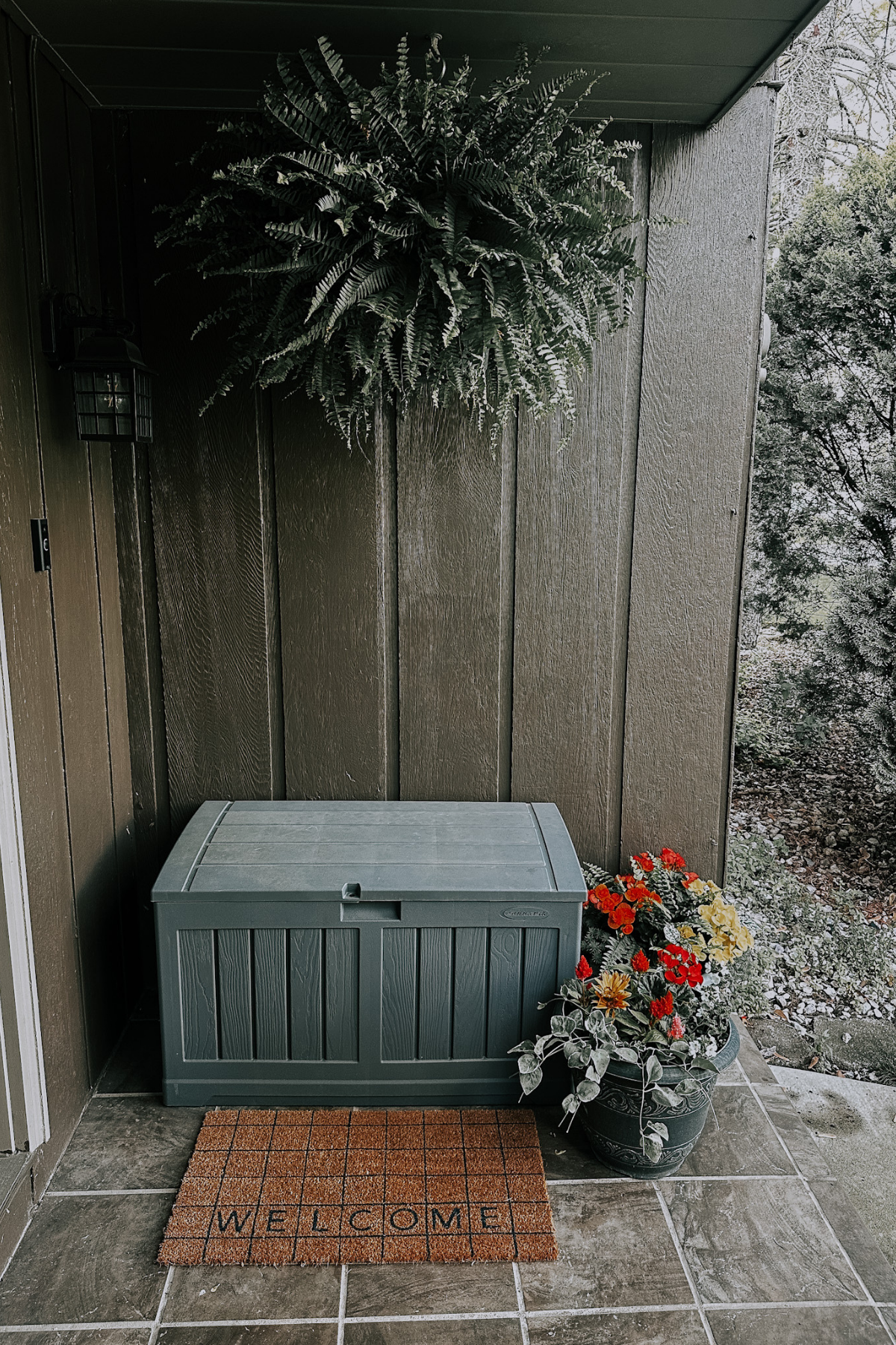 Outdoor porch area featuring a gray storage box, a welcome mat, a hanging fern plant, a potted flower arrangement with red, yellow, and white flowers, and a wall-mounted lantern light fixture.