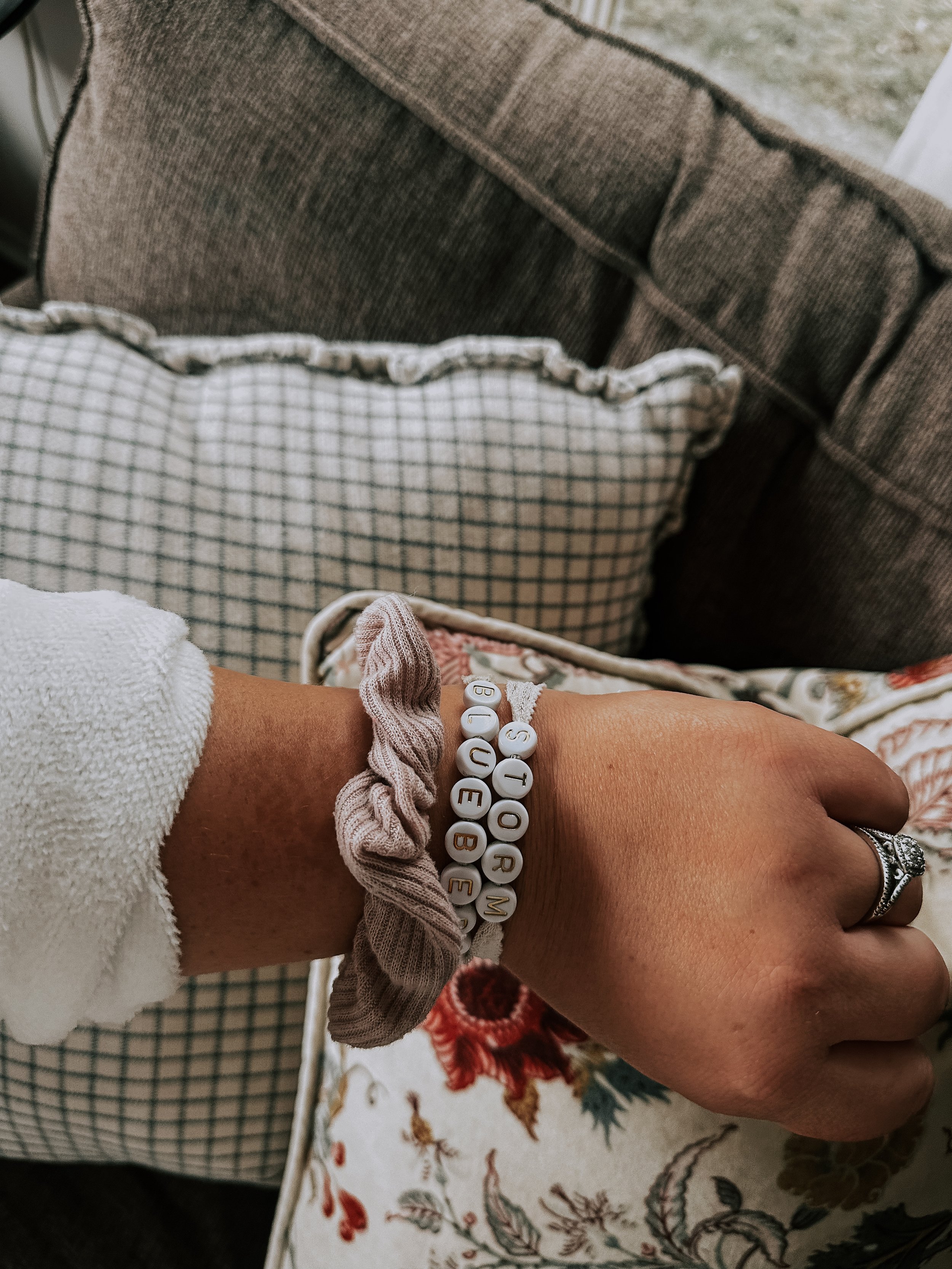 A person's hand is resting on their lap, wearing a ring and two beaded bracelets with letters on them that spell "STORM,
