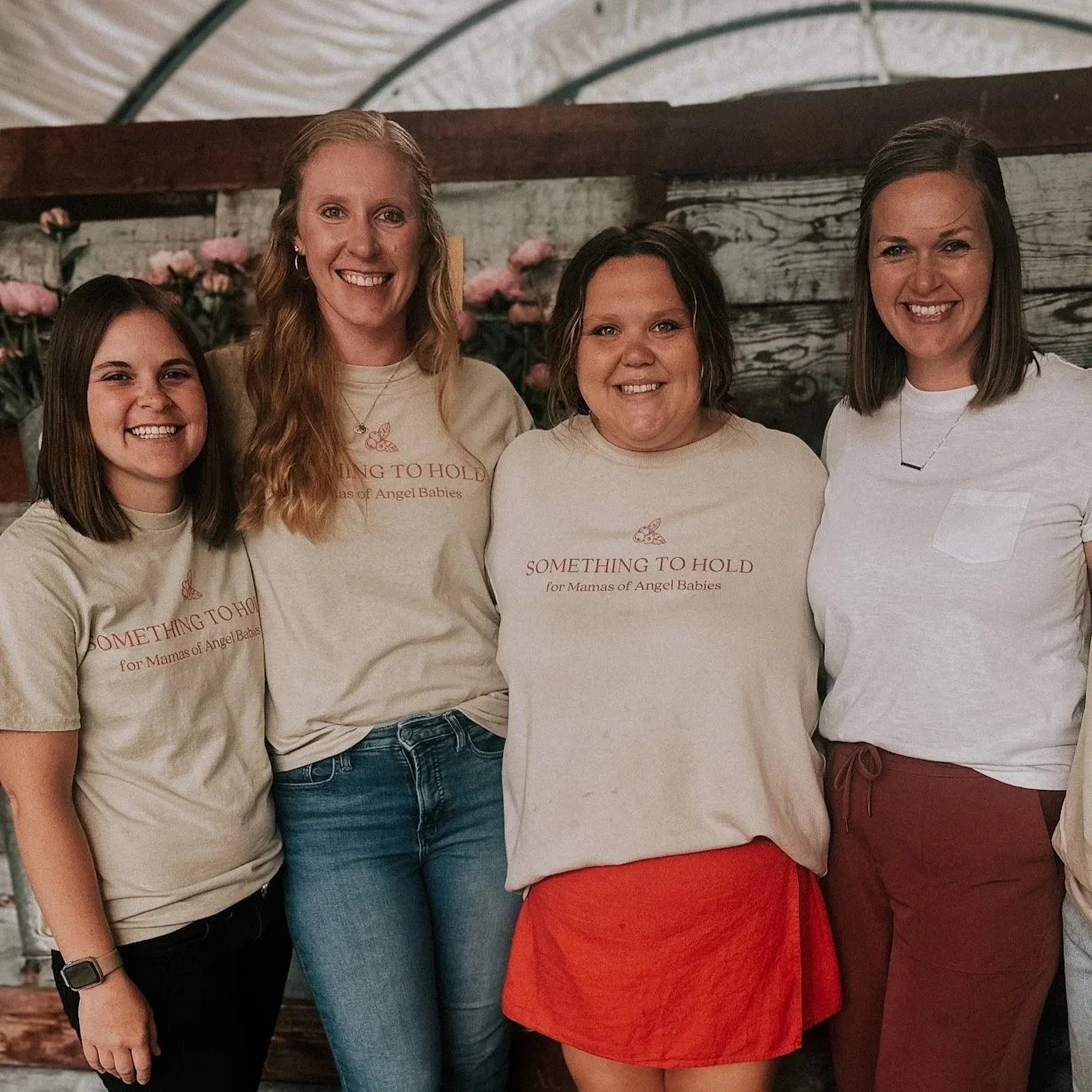 Four women standing together indoors, smiling at the camera, wearing T-shirts with a butterfly and the text "SOMETHING TO HOLD for Mamas of Angel Babies," against a rustic wooden wall with pink flowers in the background.