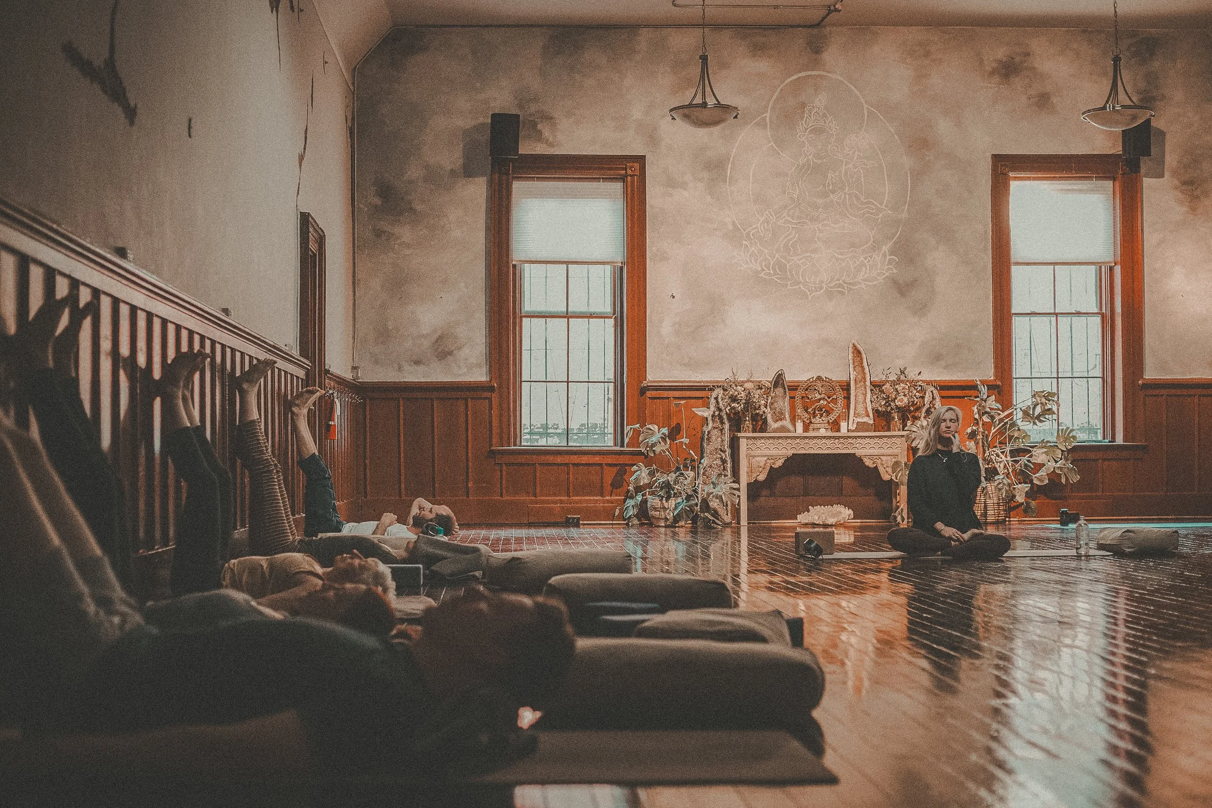 People participating in a yoga class, lying face down on mats in a dimly lit wooden indoor space.