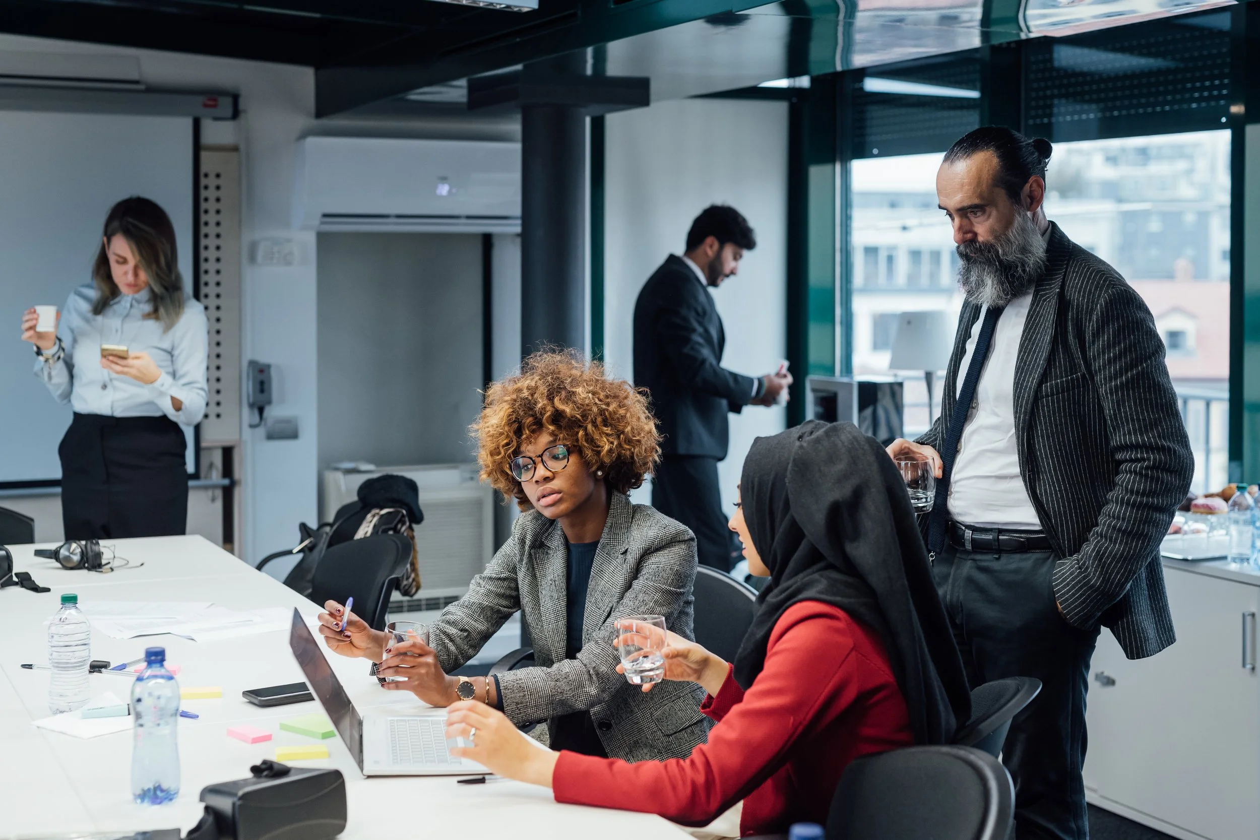 Business meeting with four ethnically diverse professionals in a modern conference room. Two women are seated at a table with laptops and water bottles, one woman in a gray blazer with glasses and curly hair, and another woman in a red blazer with a hijab. A man with a beard and striped blazer is standing, holding a glass, while another man in a suit stands in the background at the window, looking at his phone.