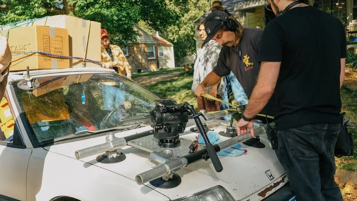 Group of people working with a camera mounted on the hood of a white car, outdoors with trees and houses in the background.