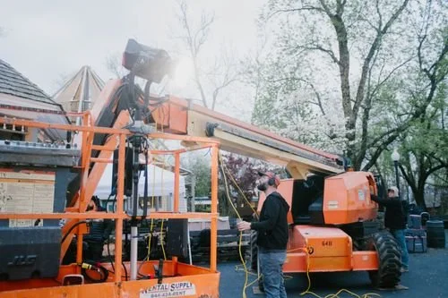 People operate an orange mobile crane outdoors among trees, with one person adjusting equipment and another nearby.