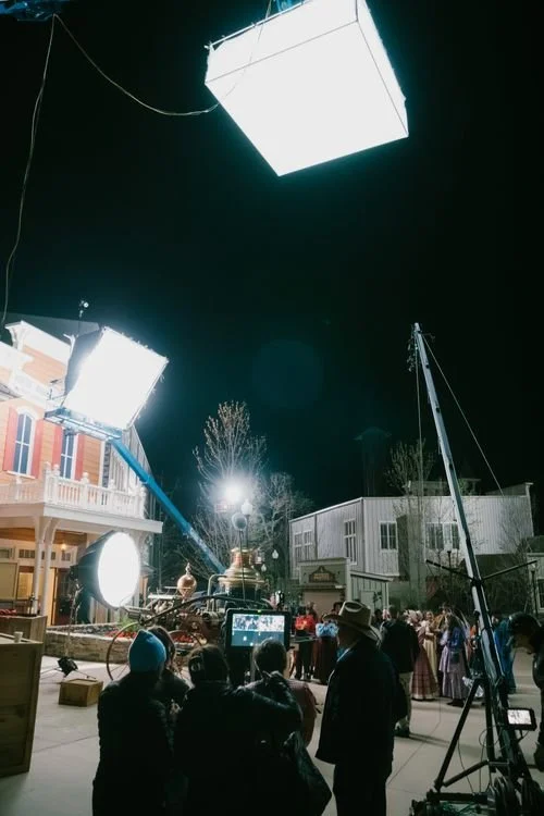 Film crew filming on set at night with lighting equipment and a crowd of people.