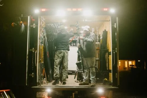 Two men working inside a well-lit grip truck at night, surrounded by tools and equipment.