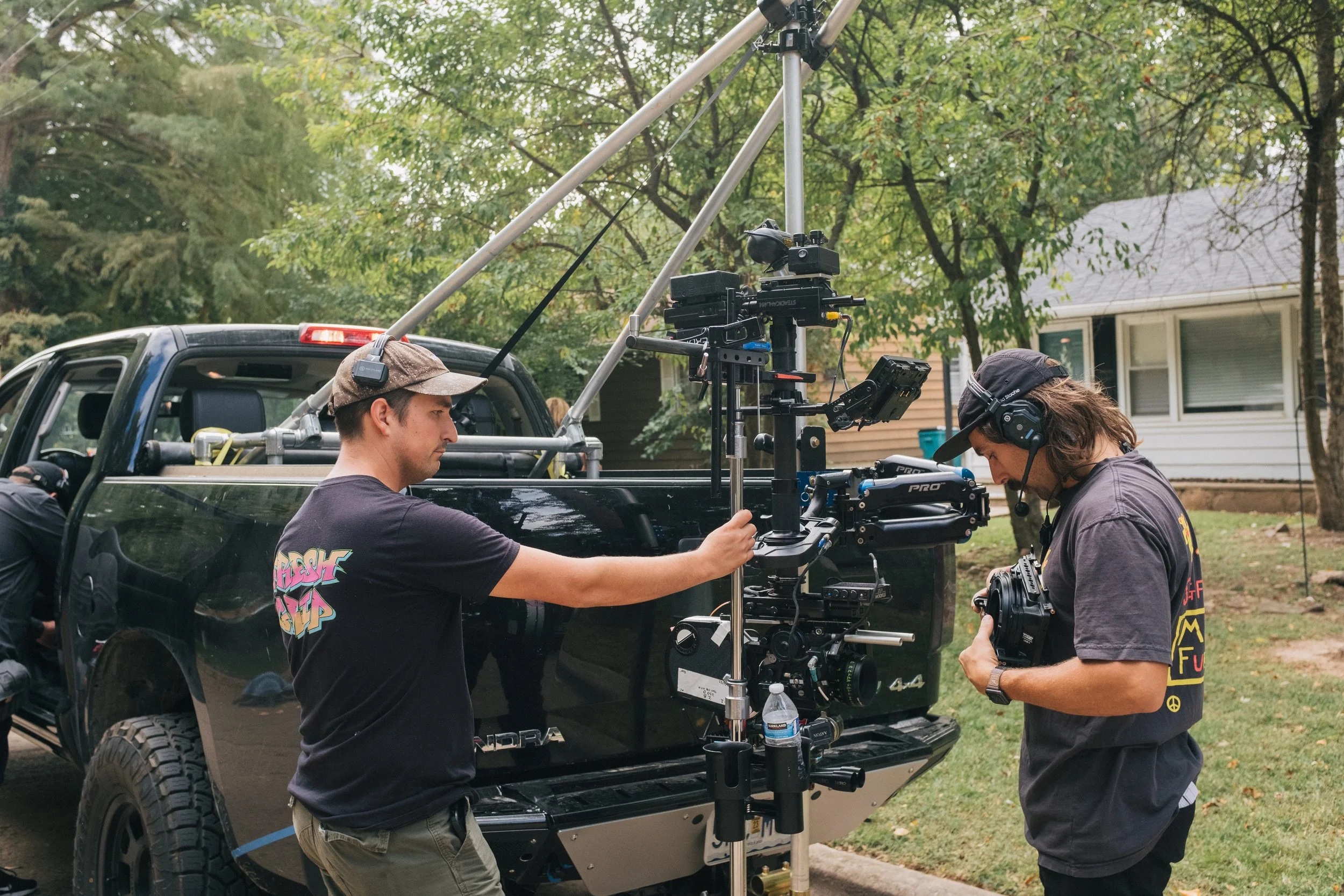 Two filmmakers working with a blackarm mounted to a black pickup truck, on a residential street with trees and houses in the background.