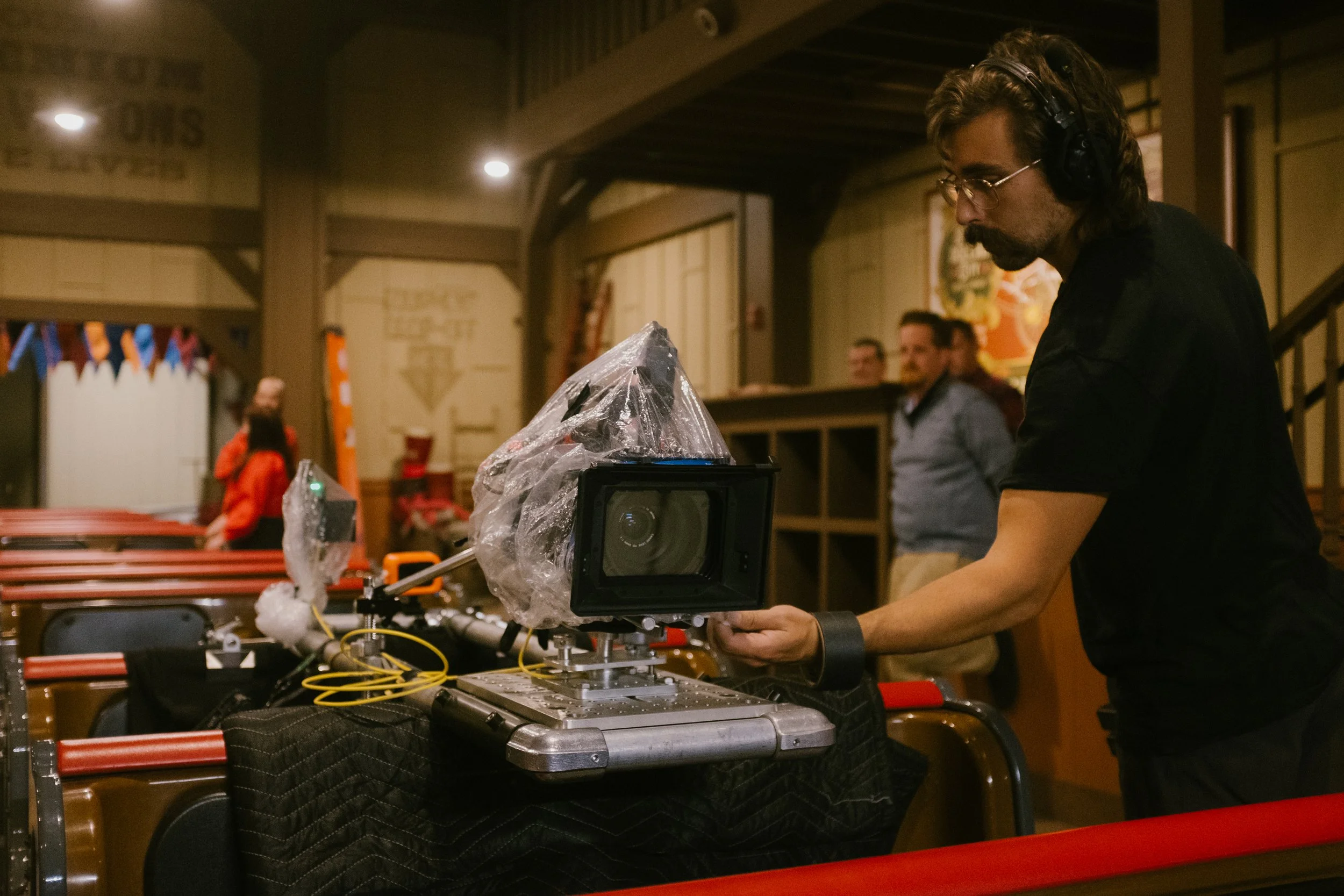 Chris Olson on set adjusting a camera rig. The camera is covered with plastic for protection, and there are people in the background inside Silver Dollar City's Fire in the Hole attraction.