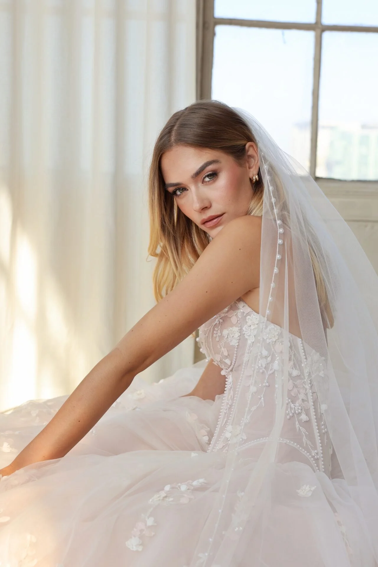 A young woman in a wedding dress with a veil, lace details, wearing a veil with pearl accents, sits near a window with cream-colored curtains in a well-lit room.