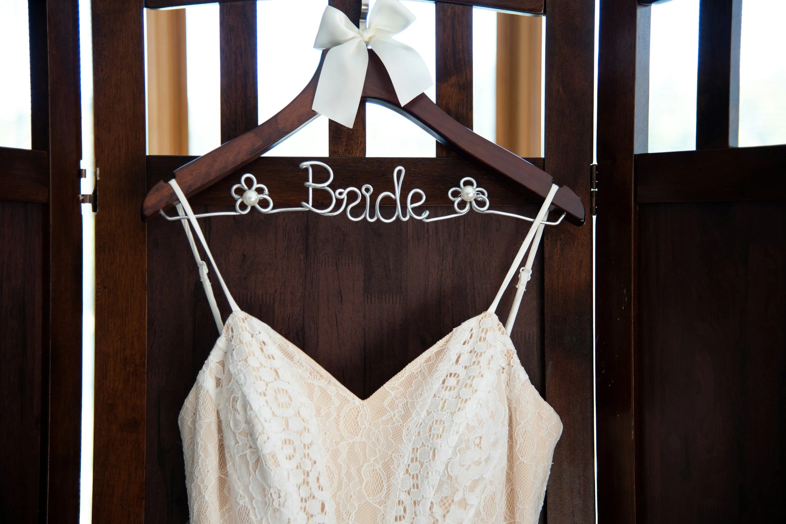 A bridal dress hanging on a wooden hanger with a decorative sign spelling 'bride' in wire, adorned with small flower accents, attached to a dark wooden background.