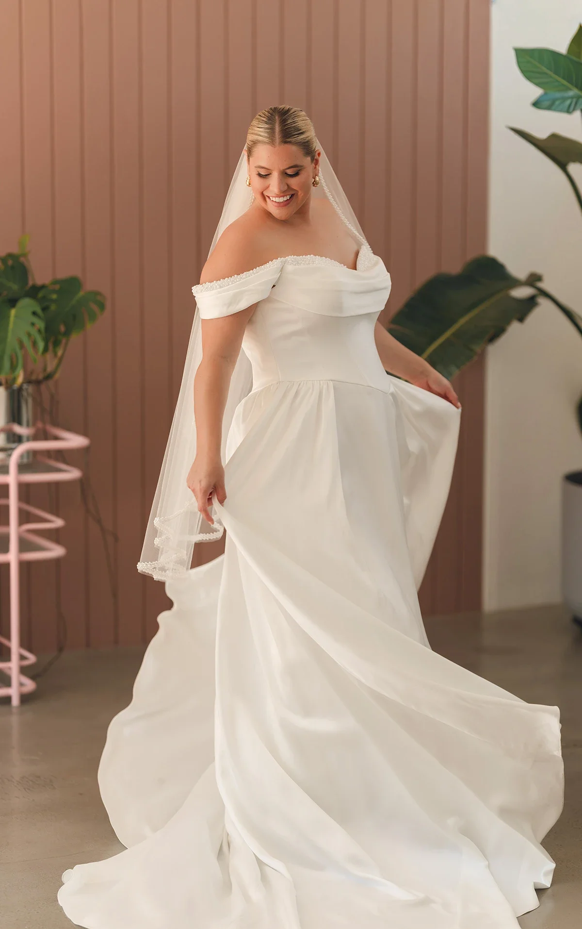 A woman in a white wedding dress, smiling and holding the skirt of her dress, standing indoors with a pink wall and green plants in the background.
