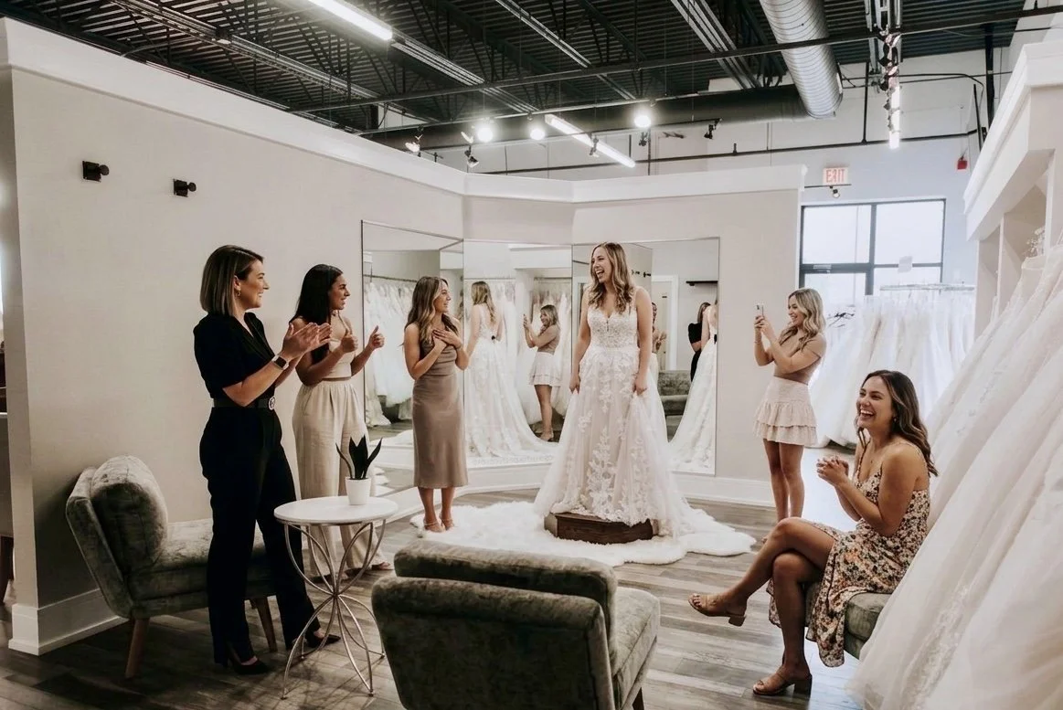 A group of women in a bridal boutique trying on wedding dresses. The bride-to-be is standing on a small platform, trying on a lace wedding gown. The friends are smiling, clapping, and taking photos.