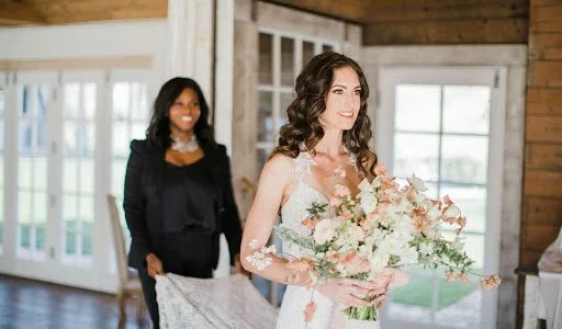 A bride holding a bouquet of flowers at her wedding, with a woman in the background in a rustic indoor setting.