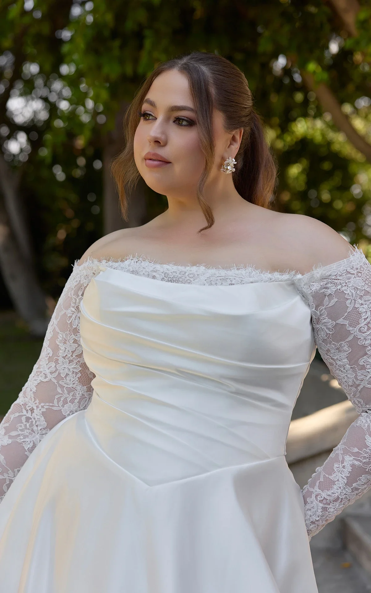 A woman wearing a strapless white wedding dress with lace long sleeves, standing outdoors with greenery behind her.