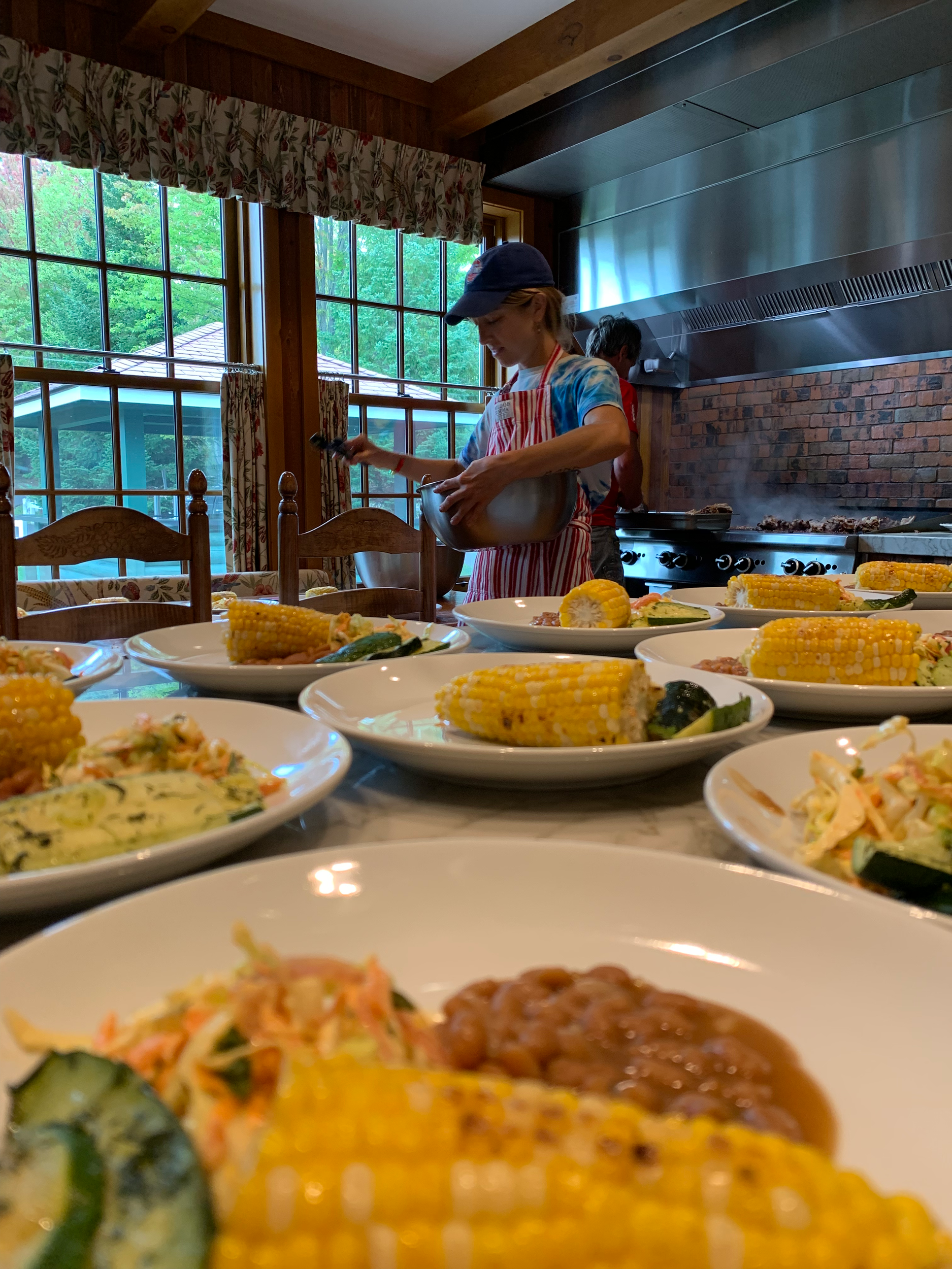People preparing food in a kitchen with a large window, various dishes on a table including corn on the cob and a salad.