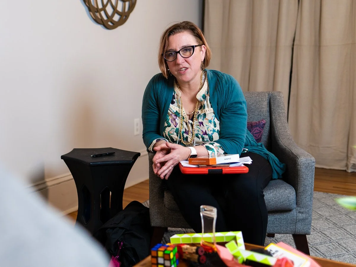 A woman with glasses and earrings, wearing a colorful blouse and teal cardigan, sitting on a gray armchair with a purple pillow, in a room with curtains and decorative wall art. There is a small black side table next to her, and a table in front with