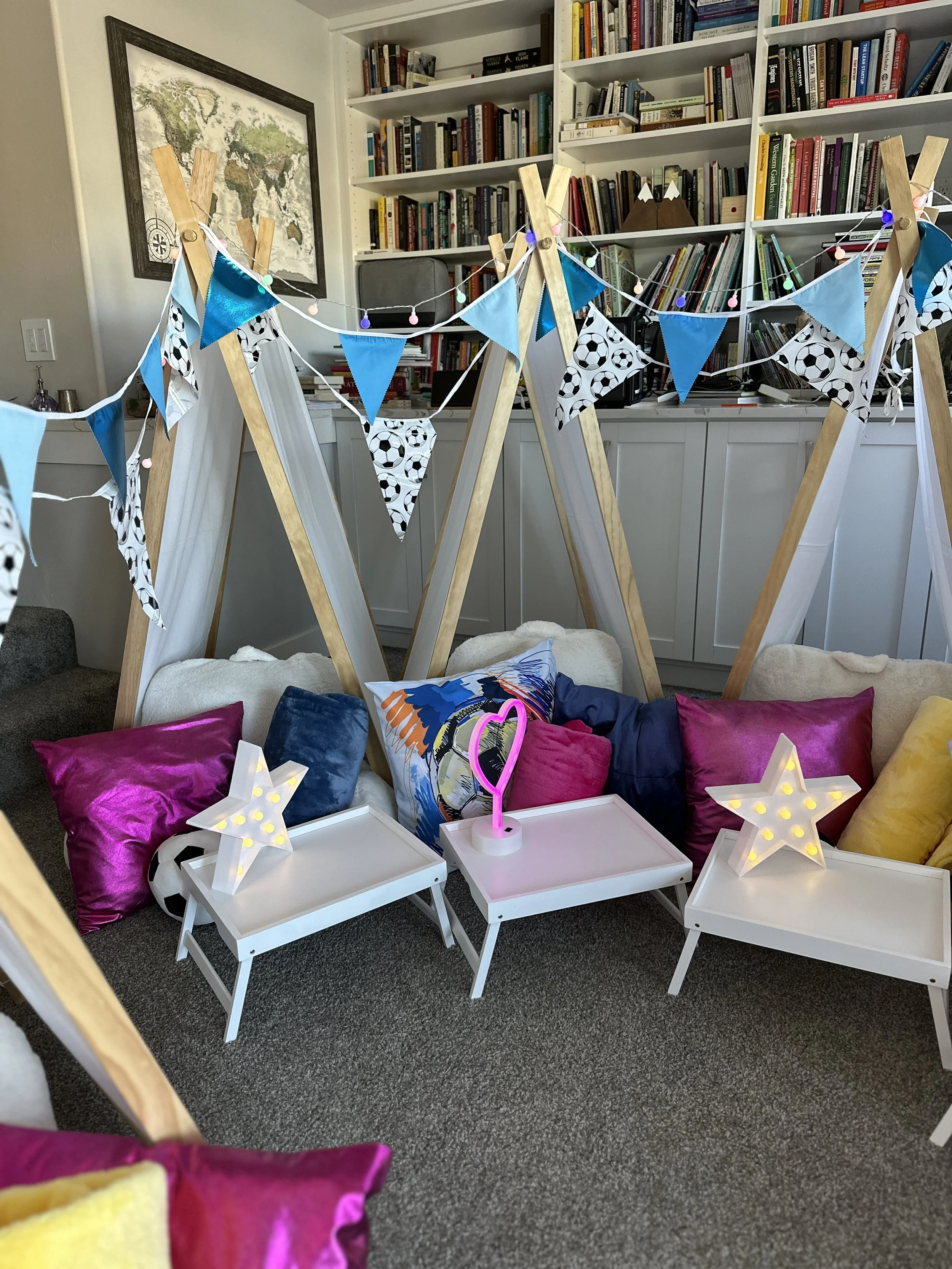 Indoor party setup with a cozy corner featuring colorful pillows, small white tables with star-shaped lamps, a neon heart light, and a teepee-like tent decorated with blue and soccer-themed bunting. A bookshelf and world map are visible in the background.