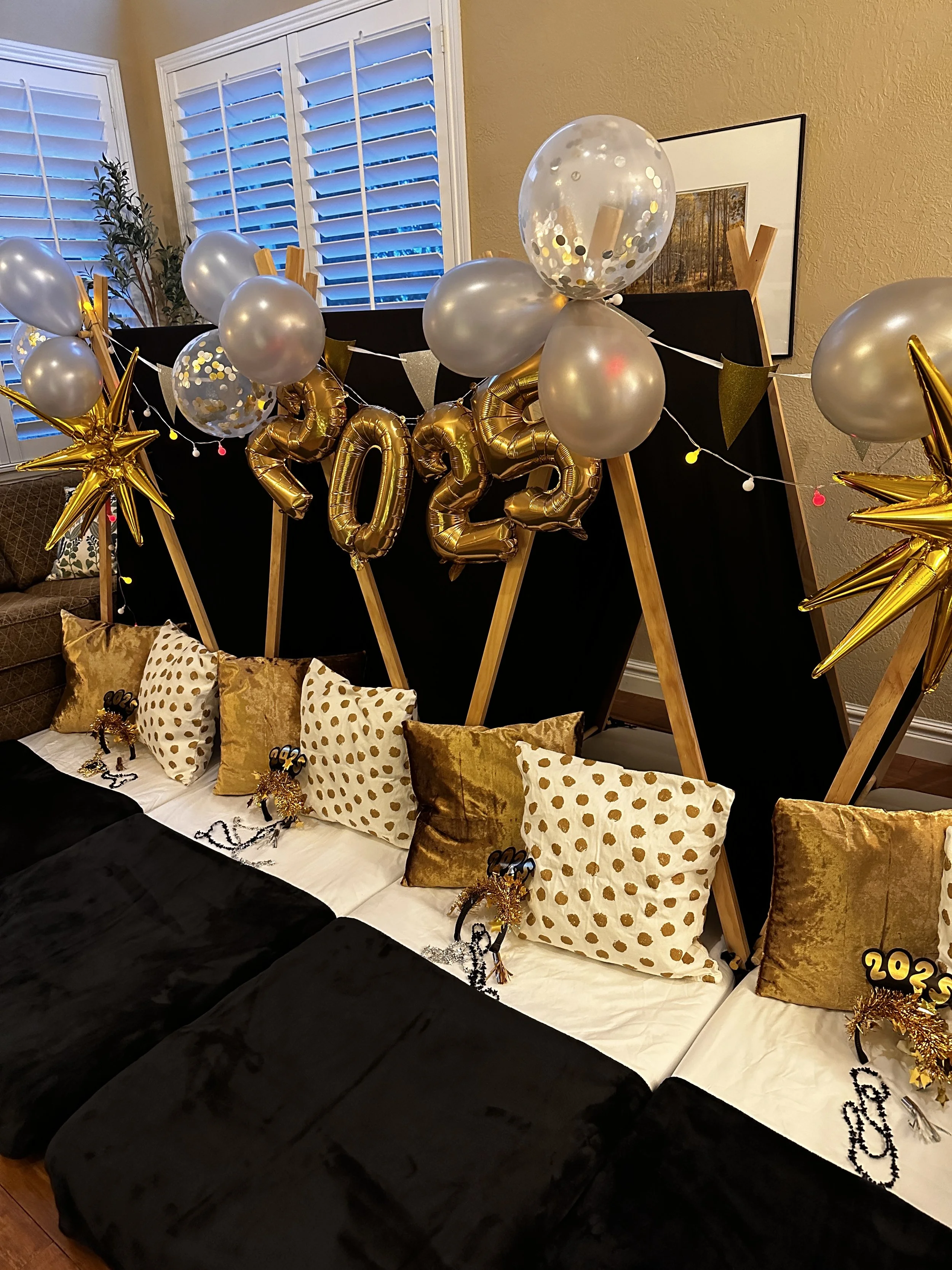 Decorative New Year's Eve display featuring balloons and balloons spelling 2023, with gold and silver color theme, gold star and polka dot pillows, and small party favors on a couch with a black backdrop.