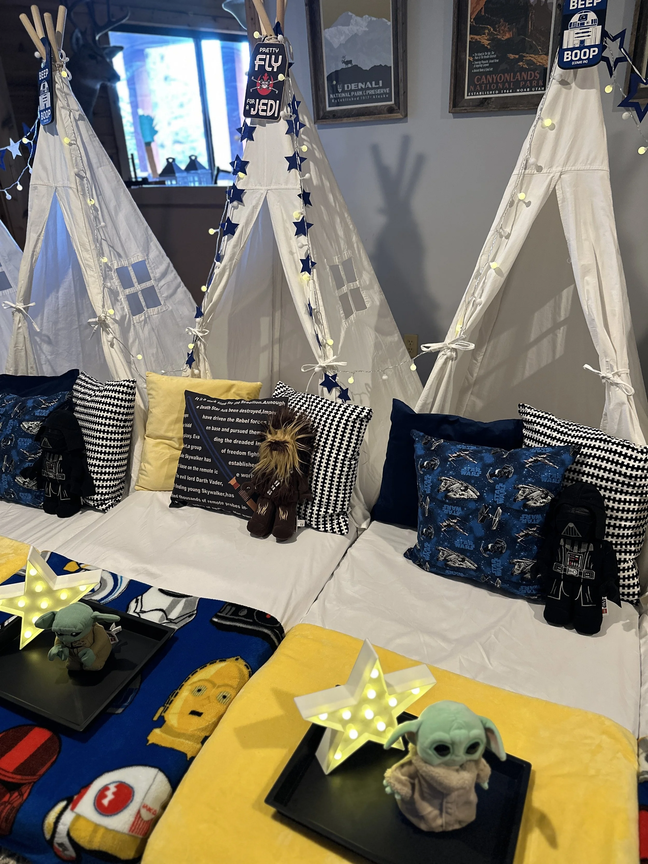 Star Wars themed bedroom with white teepees decorated with blue star garlands and string lights. Beds are covered with Star Wars pillows and plush toys include Baby Yoda, Darth Vader, and other characters. Small trays with illuminated star decorations and plush toys are on the beds.