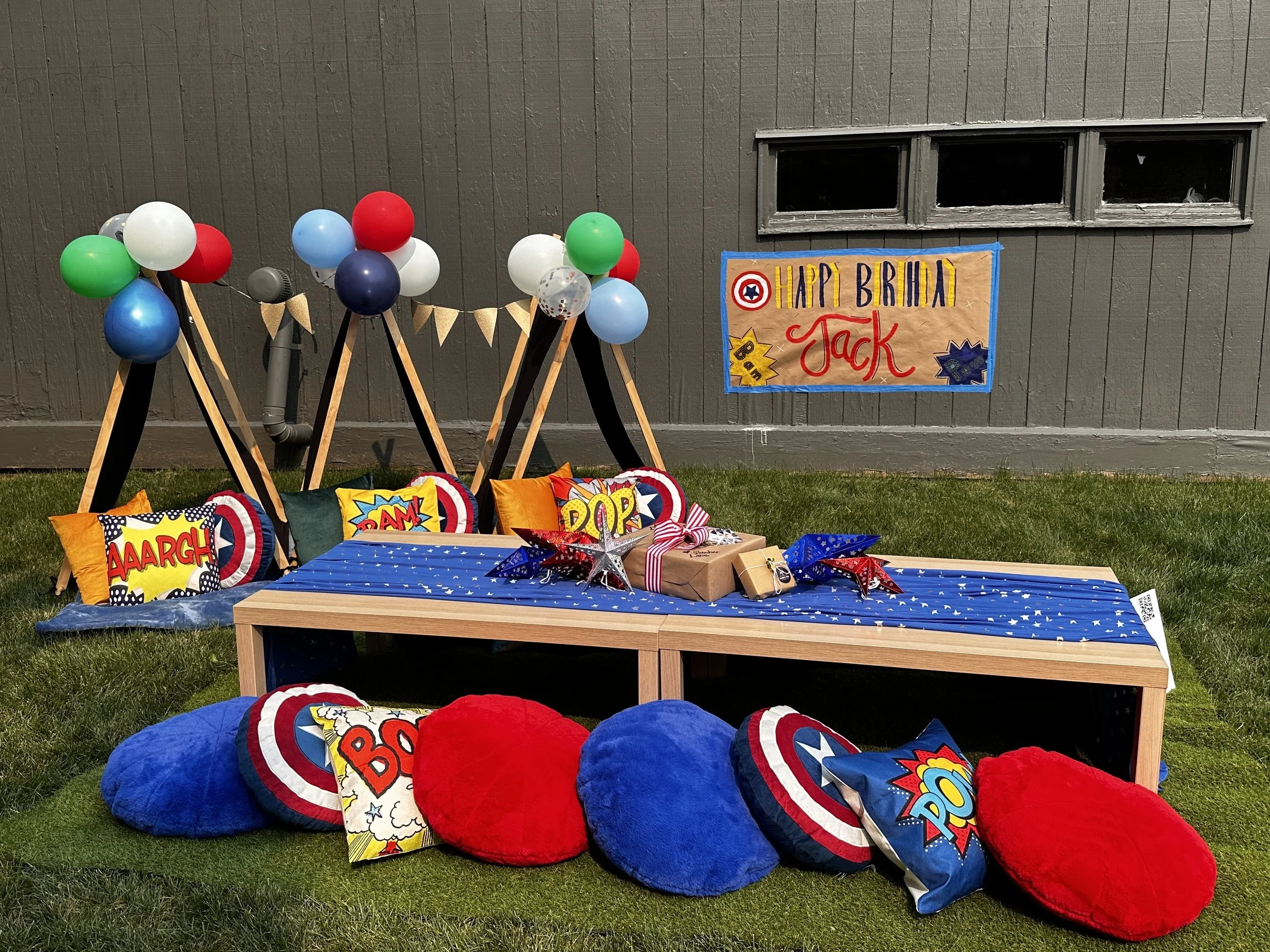 Superhero-themed birthday party setup with blue, red, green, and white balloons, comic book style pillows with words like 'AARGH,' 'BAM,' 'POW,' and 'BOOM,' a table with starry blue cloth, wrapped gifts, and a colorful sign saying 'Happy Brinal Jack' in front of a gray wall with narrow windows.