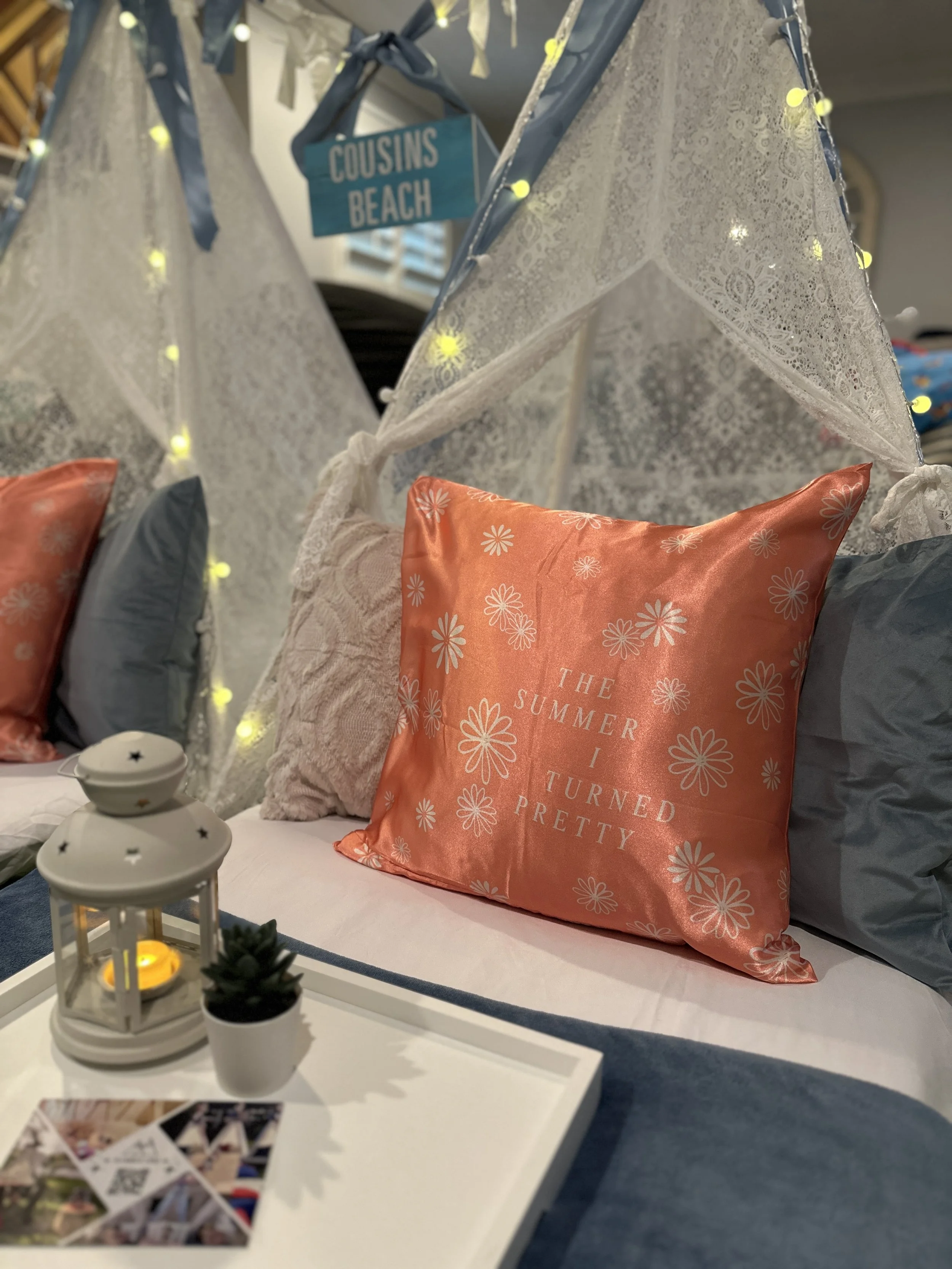 Decorative scene with a bed featuring pillows, including one with the text 'The Summer I Turned Pretty' in front of a white tray holding a small lantern and a tiny potted plant. There is a tent-like canopy with string lights and a sign that says 'Cousins Beach' hanging above.