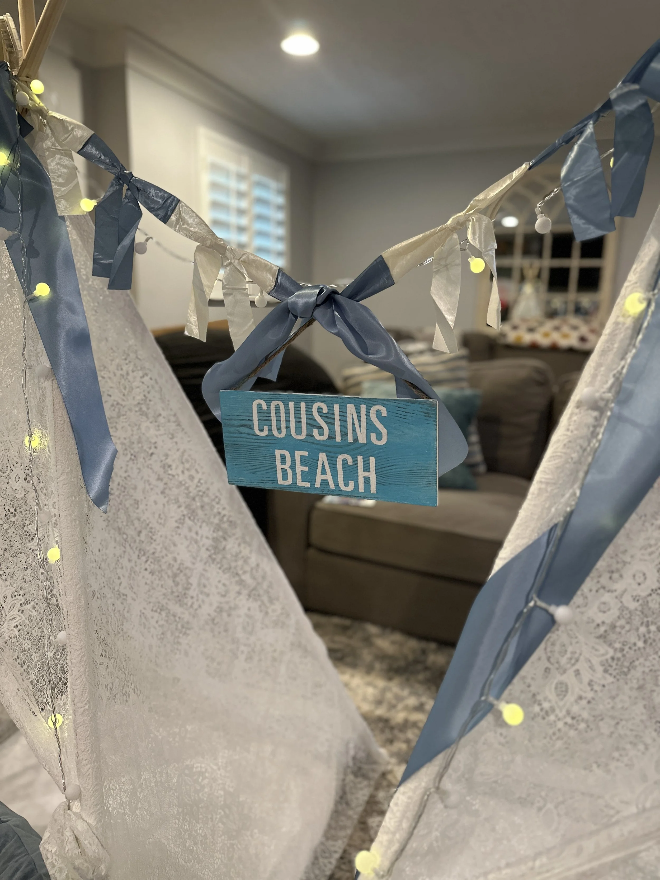 A decorative sign that says 'Cousins Beach' hanging inside a fabric teepee with lace and ribbon decorations, in a living room.