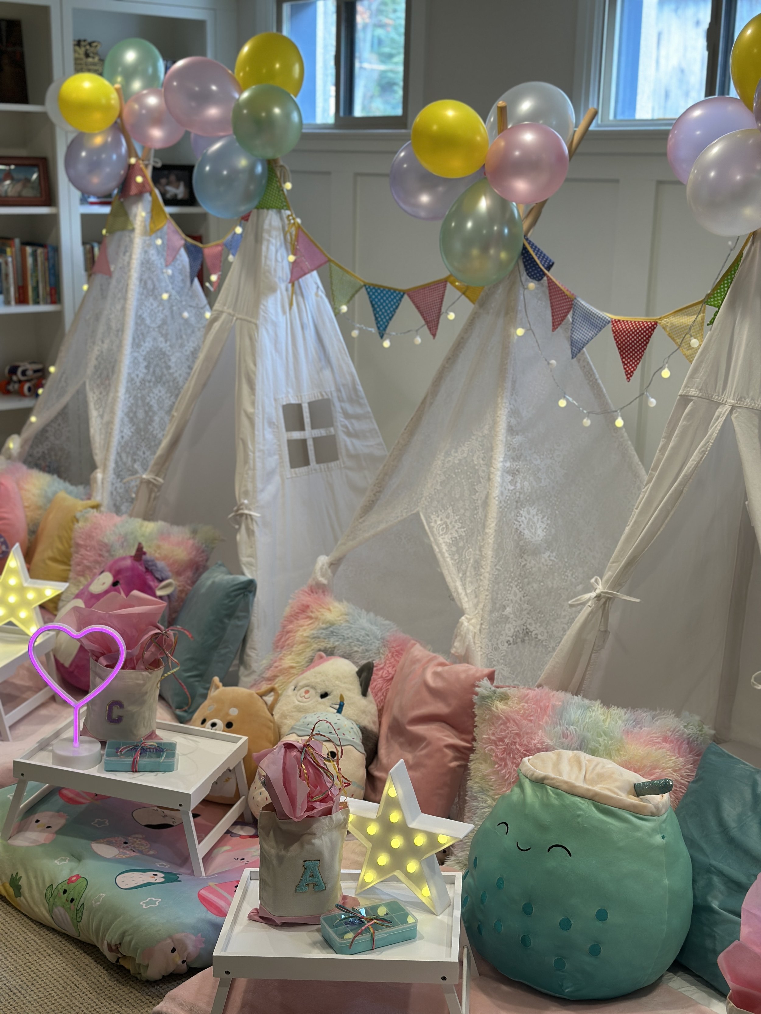 Decorated indoor space with small teepees, colorful balloons, fairy lights, plush toys, pillows, and decorative lamps for a children's play area or party.