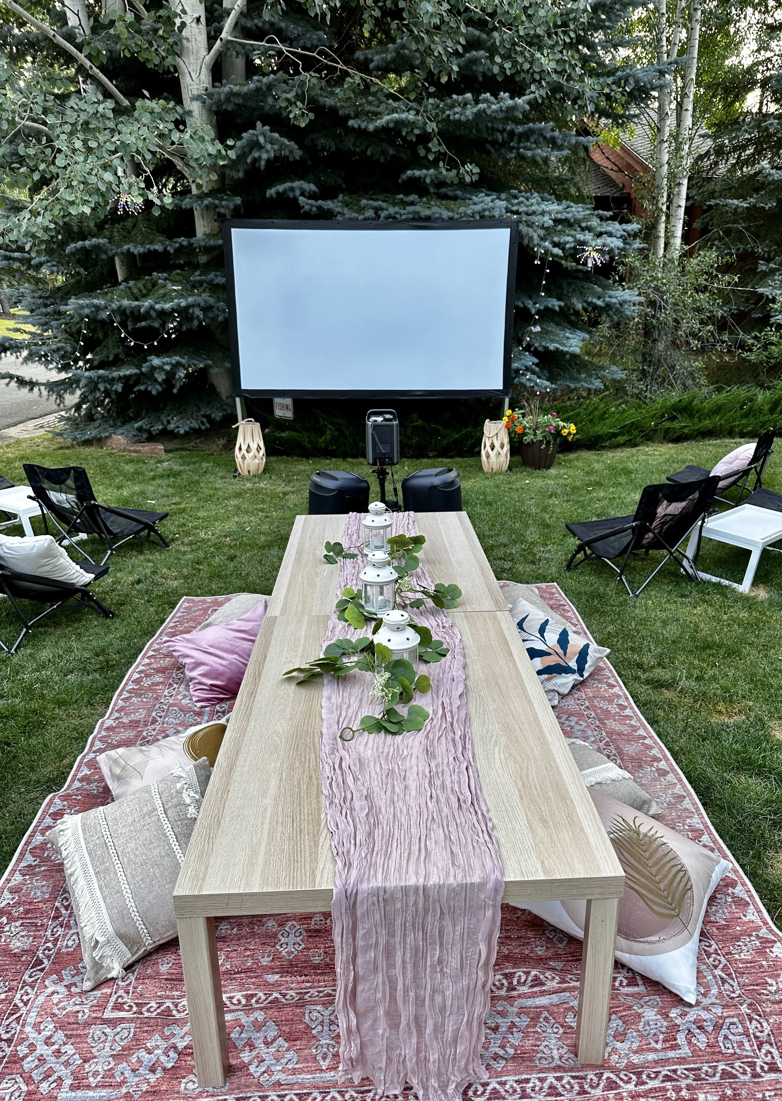 Outdoor backyard movie setup featuring a large projection screen, foldable chairs, and a dining table with pillows, lanterns, and greenery, surrounded by trees.