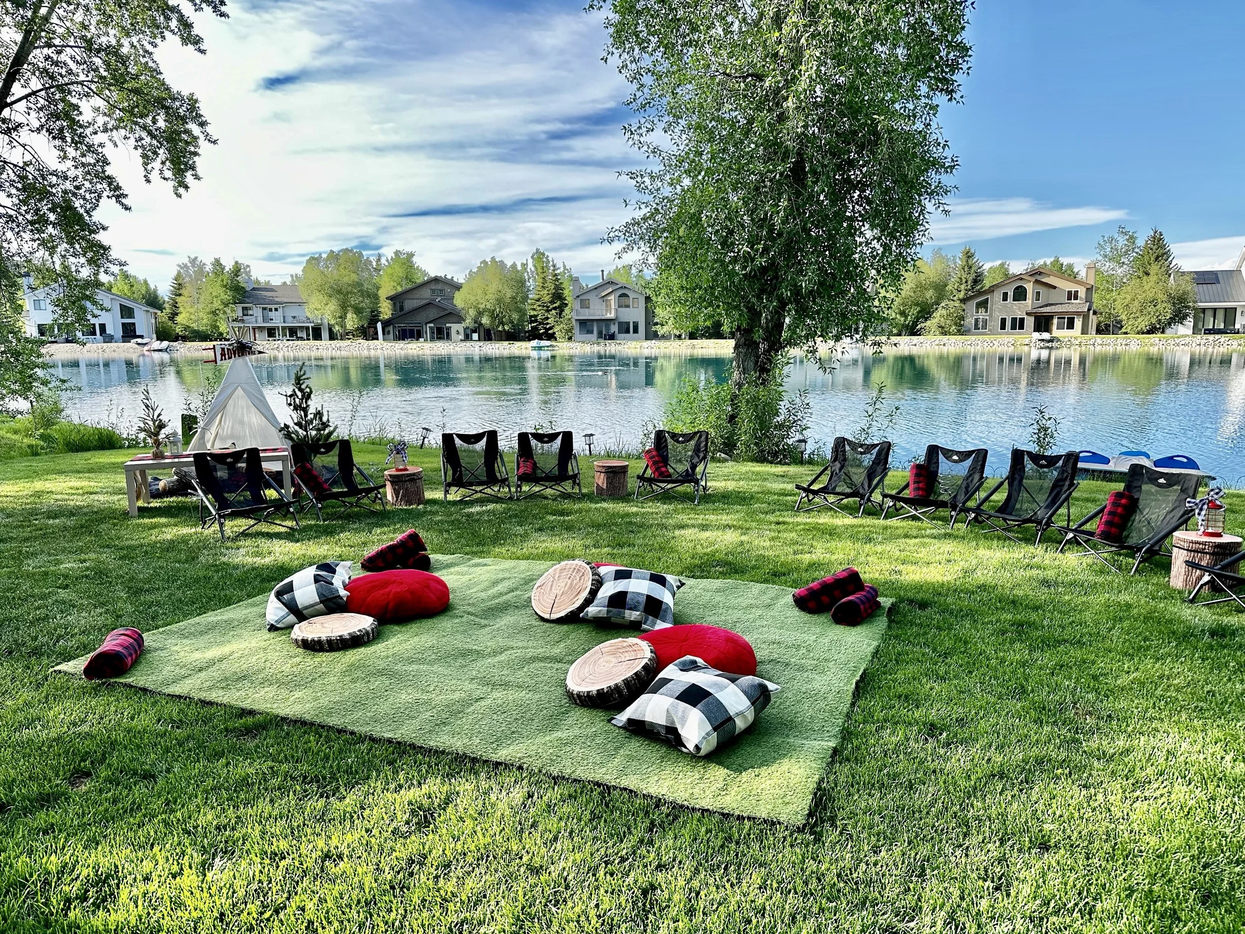 A lakeside outdoor setup with a grassy area featuring cushions and logs on a green rug, surrounded by lounge chairs and small tables, overlooking calm lake water with houses and trees in the background on a partly cloudy sky day.
