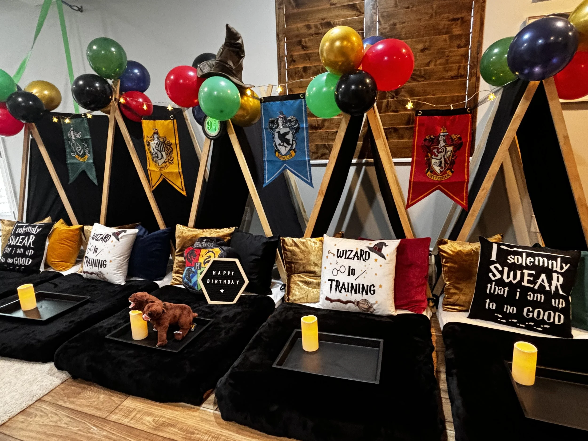 Interior decorated for a Harry Potter-themed birthday party or event, featuring banners with house crests, colorful balloons, and cushions with Harry Potter-related sayings, with a small stuffed animal on the black sofa.