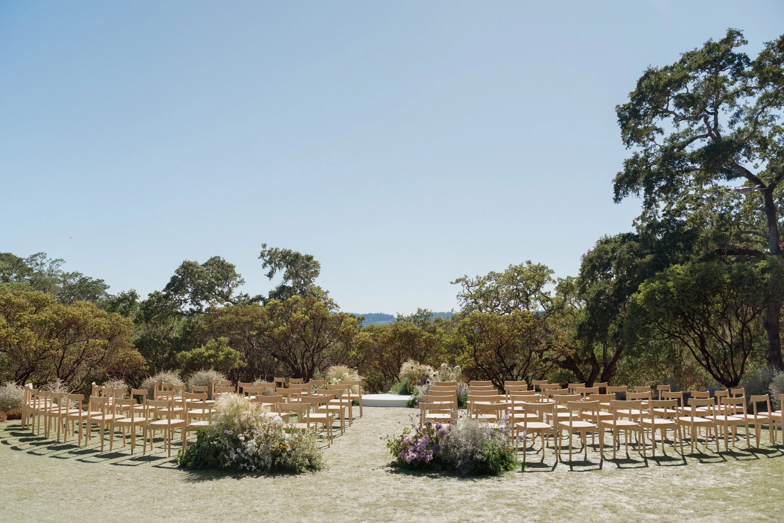 Outdoor wedding ceremony setup with rows of chairs, flower arrangements, and a platform on a grassy field under a clear sky, surrounded by trees.